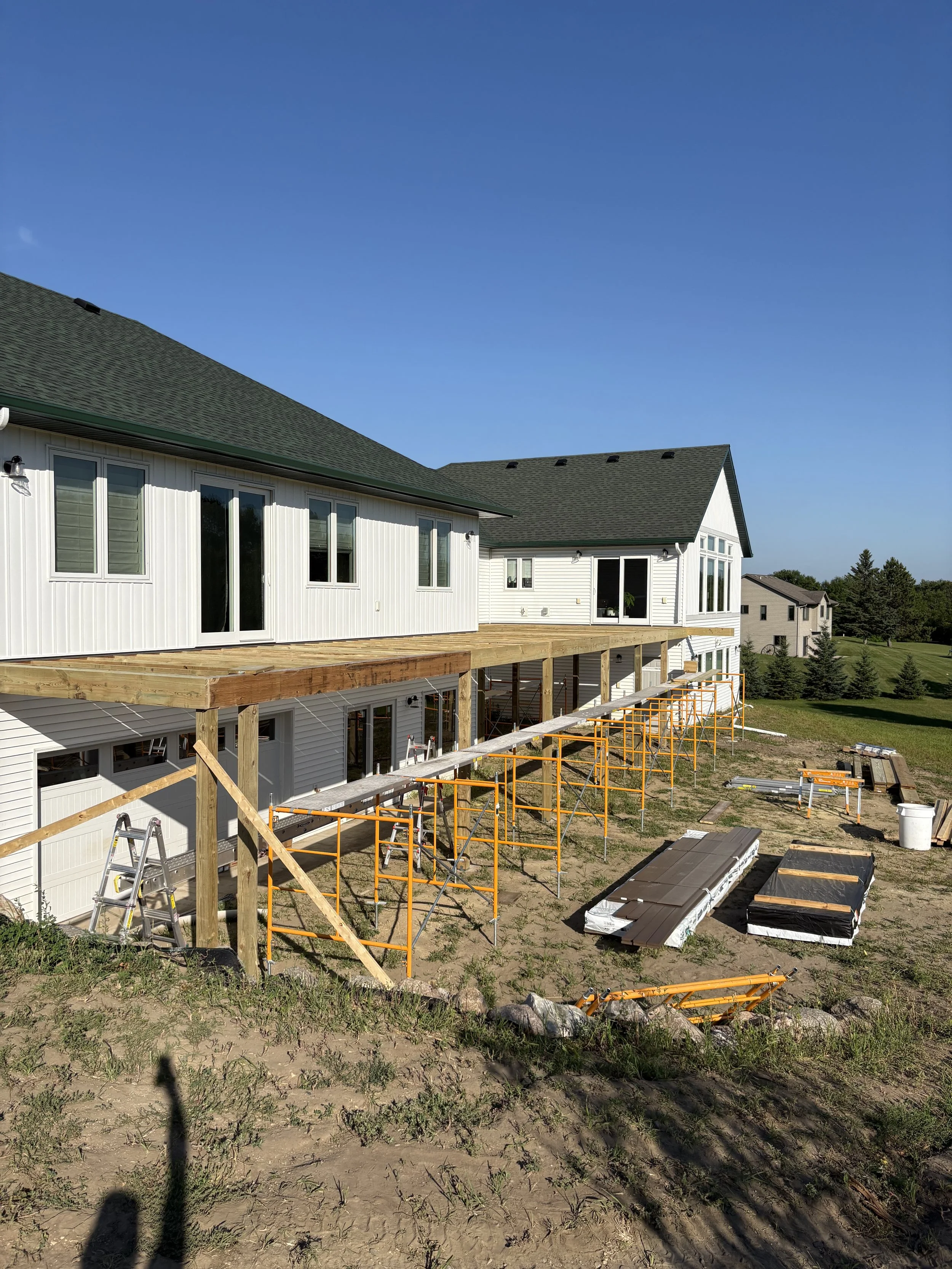 The backside of a white house under construction with scaffolding and a wooden deck, with neighboring houses and green lawn in the background.
