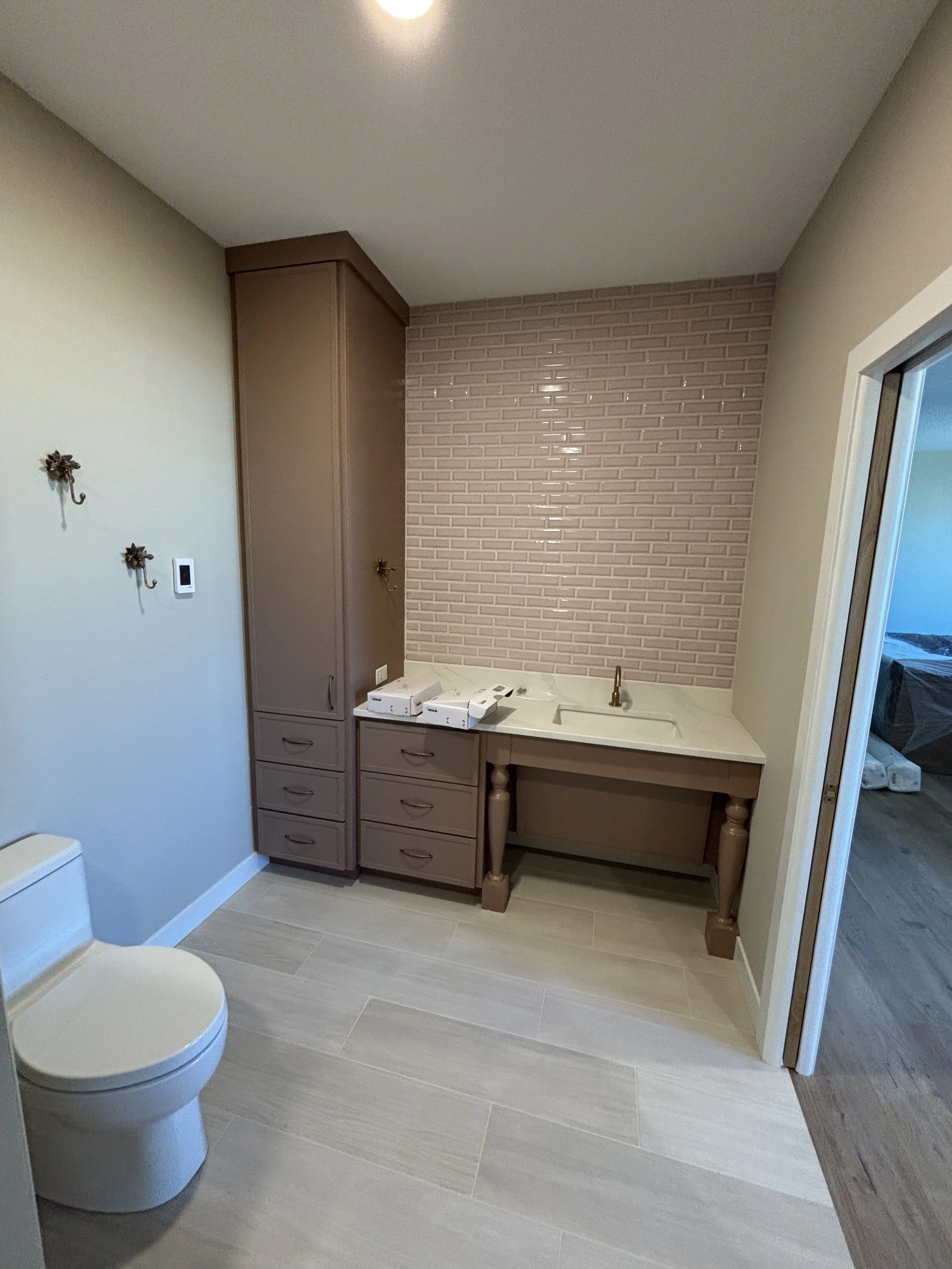 A partial view of a bathroom with a toilet in the foreground, a small cabinet, a built-in sink, a pink brick accent wall, and a doorway leading to another room.