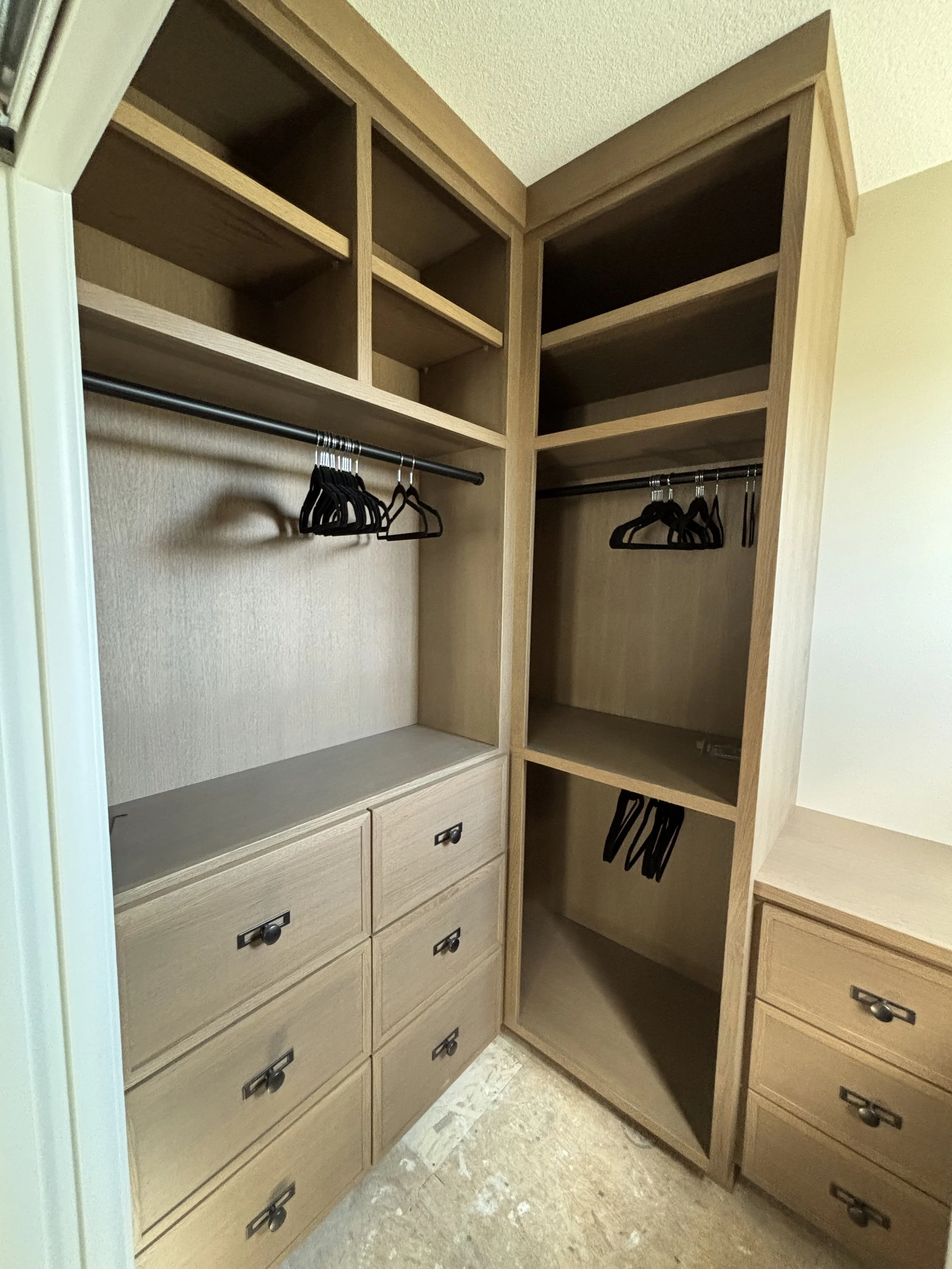 Empty wooden closet with shelves, drawers, and hanging rods, some with black hangers, in a room with beige walls and flooring.