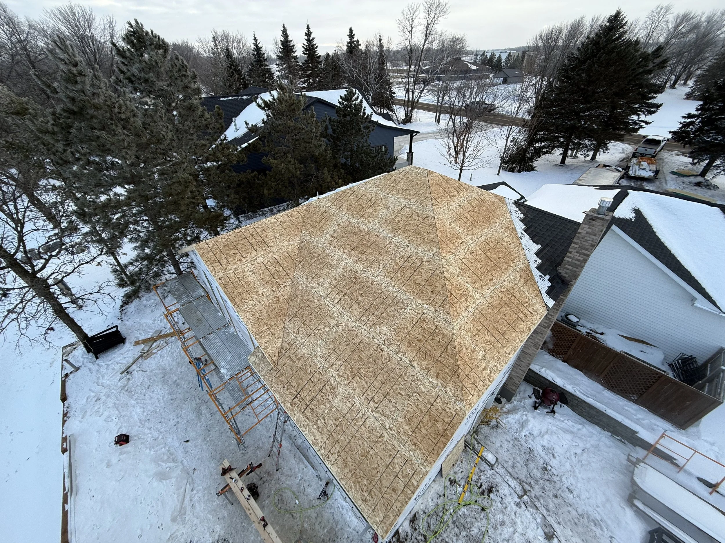 Aerial view of a house under construction with a plywood roof, surrounded by snow, with trees and neighboring houses in the background.