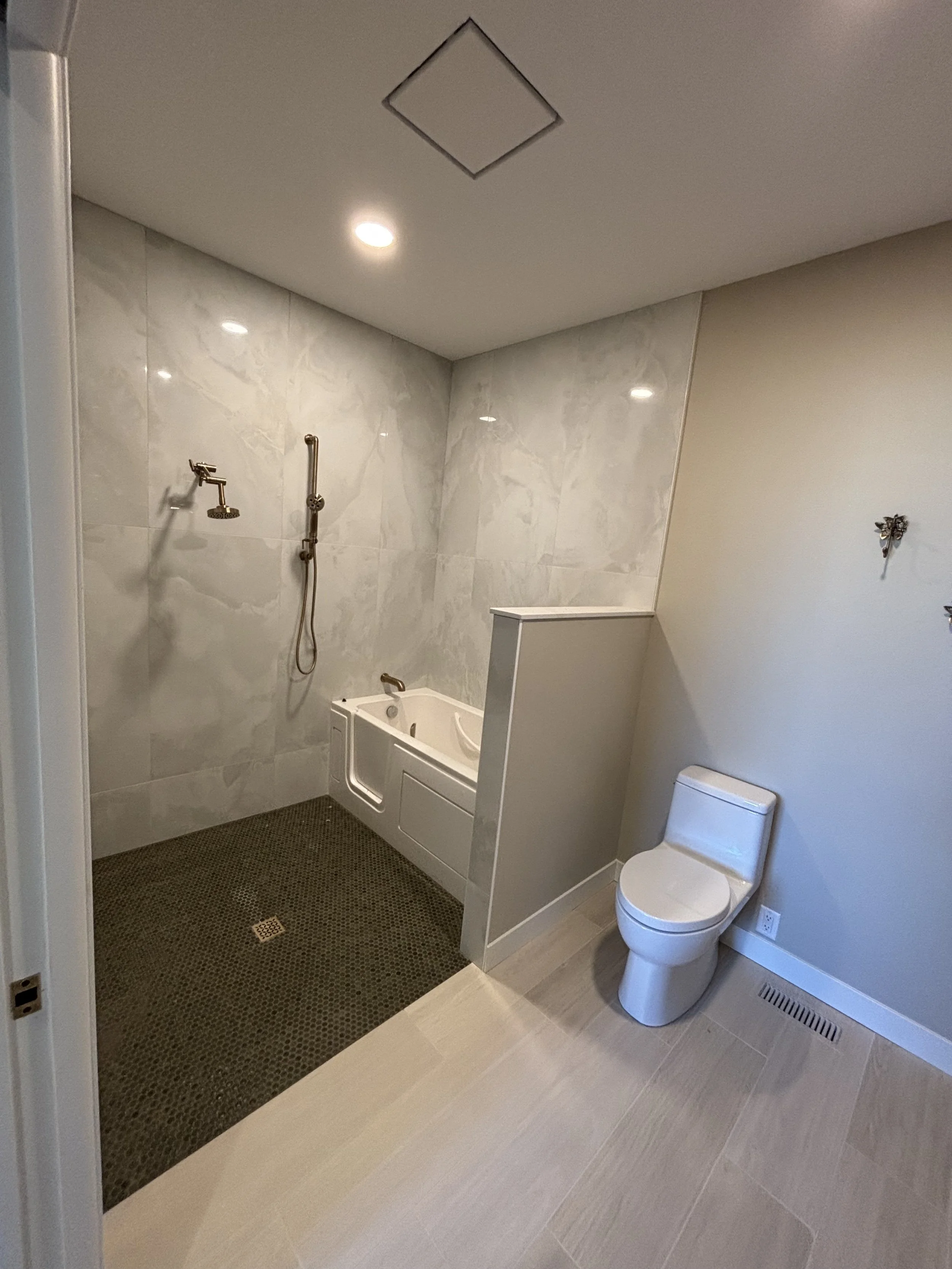 Bathroom with a walk-in shower area with a hand-held showerhead, a small bathtub, a toilet, and a beige tiled floor.