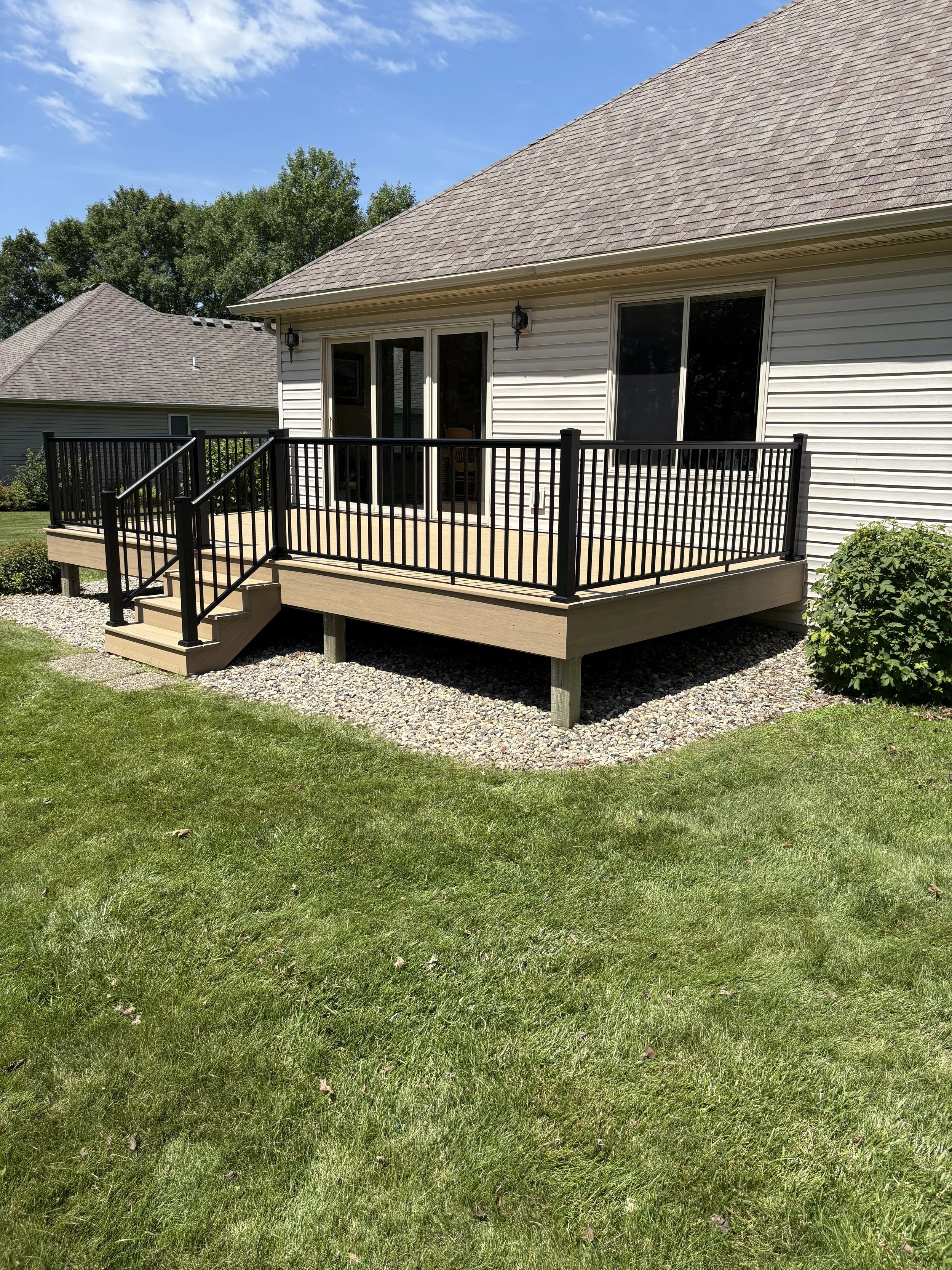 Backyard with a wooden deck, black metal railing, sliding glass door, and white vinyl siding, surrounded by green grass and trees under a blue sky.