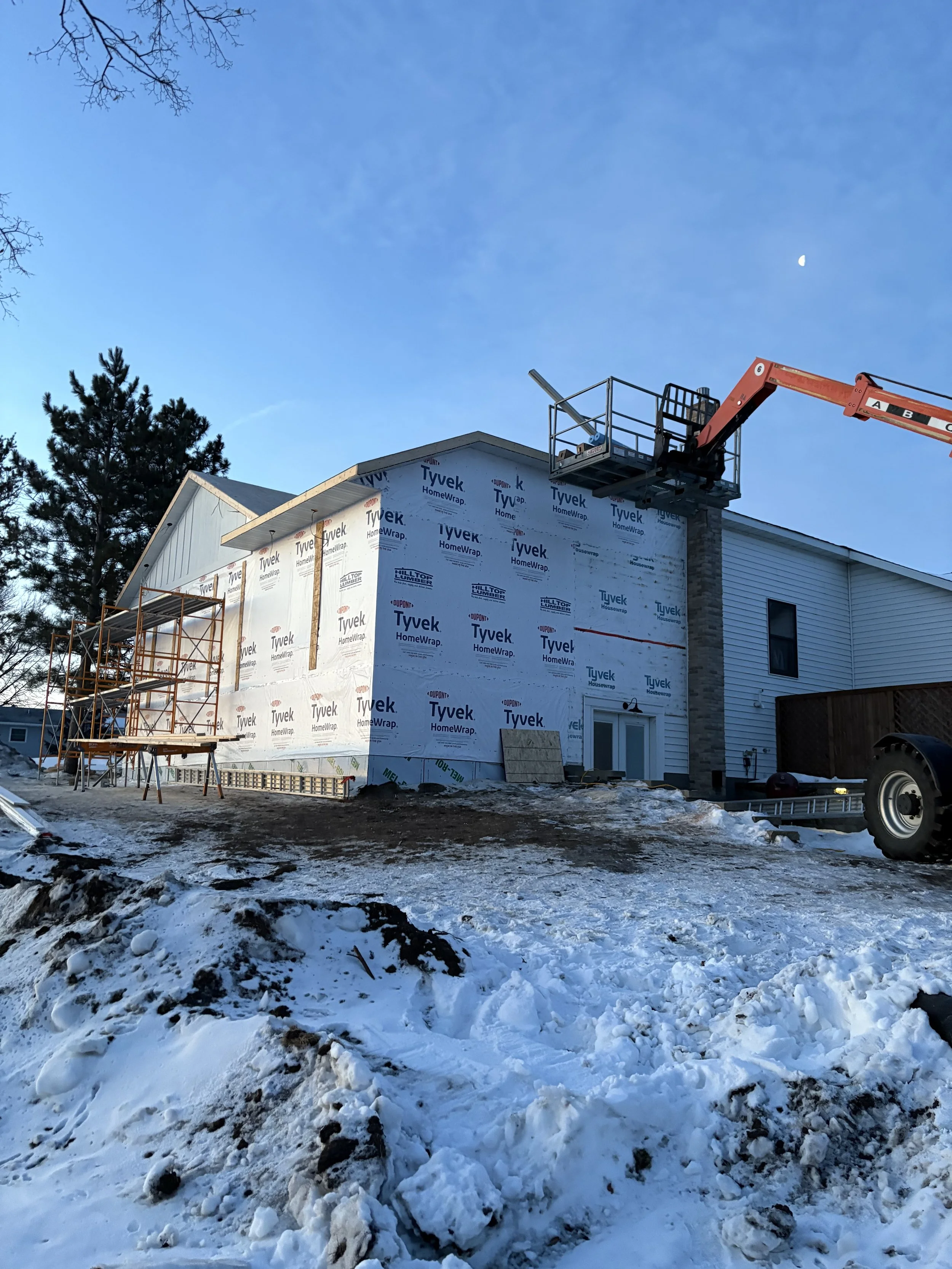 Construction site with a partially built house covered in Tyvek HomeWrap, a cherry picker lifting a worker, snow on the ground, a tree, and the moon in the evening sky.