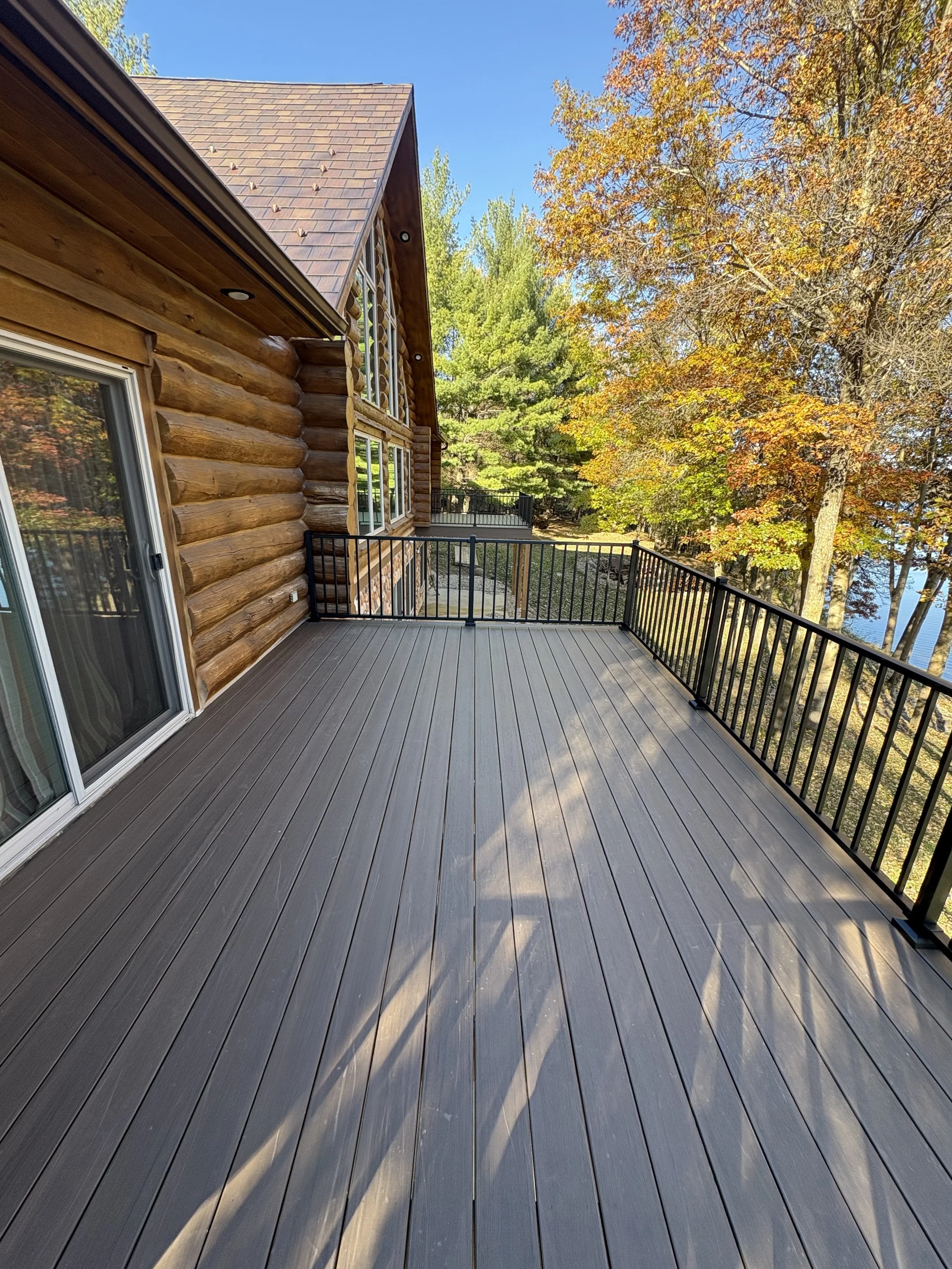 A spacious outdoor balcony with wood flooring and a black metal railing, attached to a log cabin-style house with a shingled roof, surrounded by trees with green and orange leaves, indicating fall, under a clear blue sky.