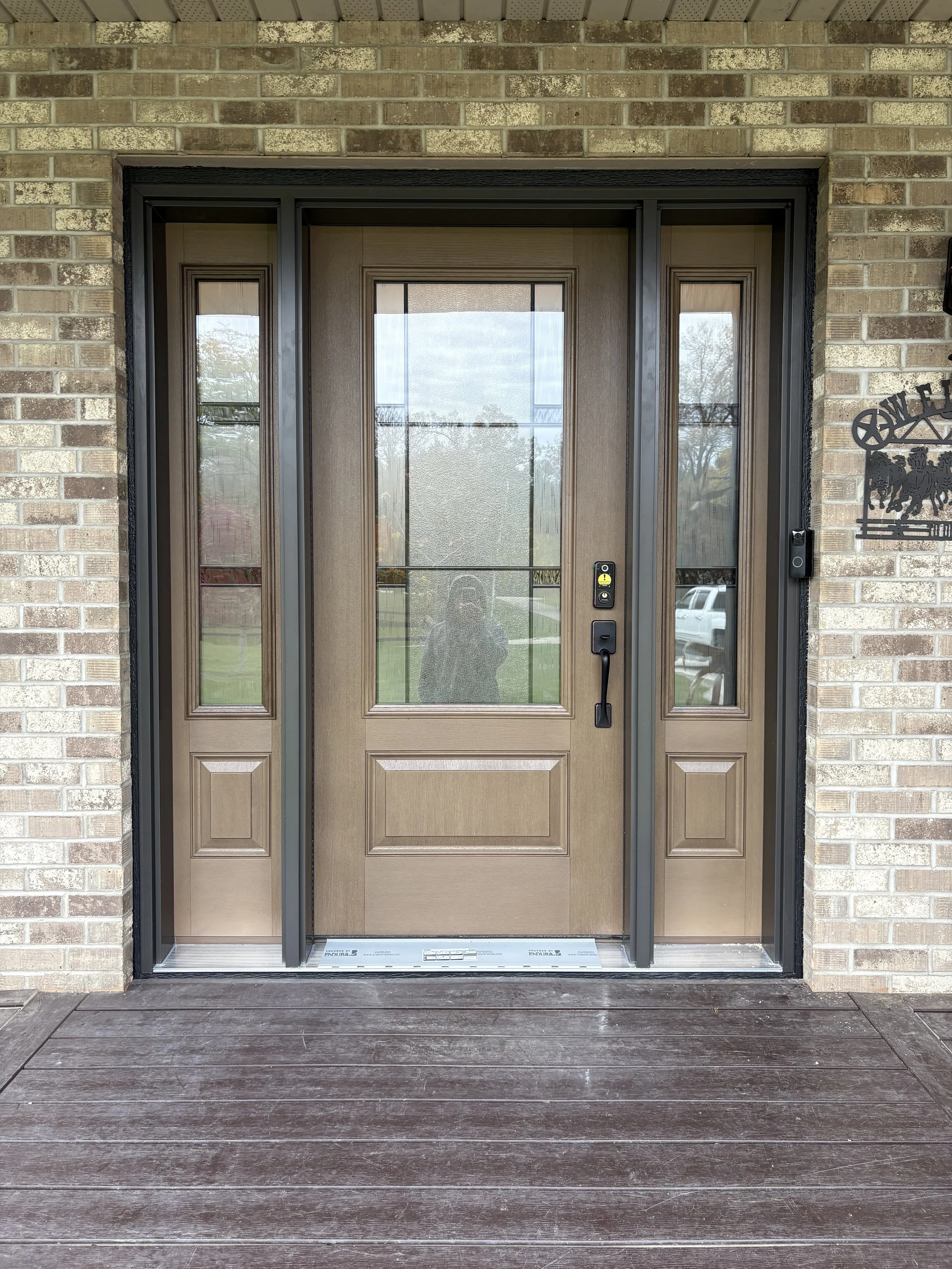 Front view of a modern wooden front door with glass panels on the sides and a brick wall surrounding it, leading to a porch with a wooden deck.