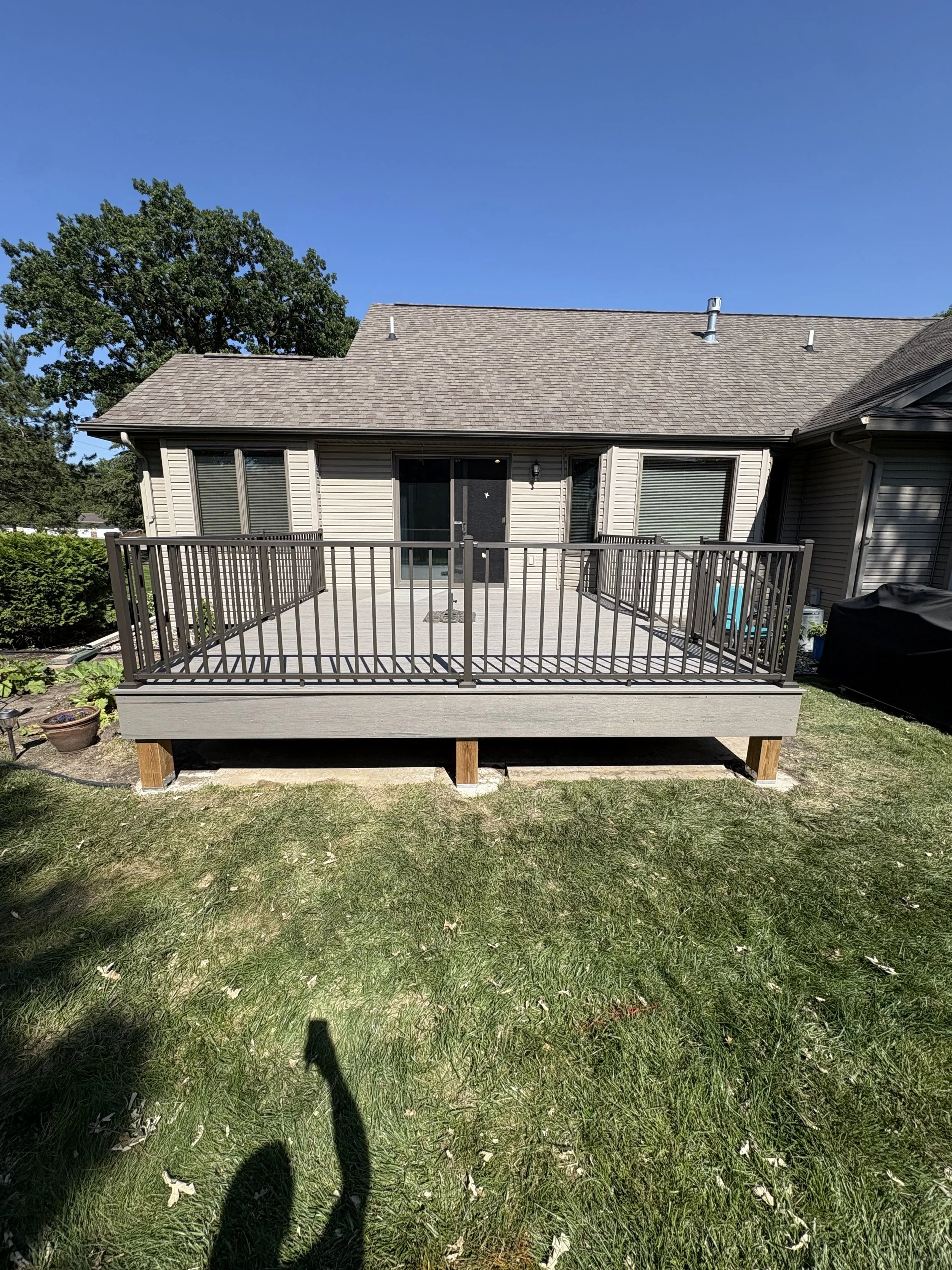 A backyard deck attached to a house with beige siding. The deck has metal railings and a door leading inside. The yard has grass and small plants, with a large tree in the background and a clear blue sky overhead.