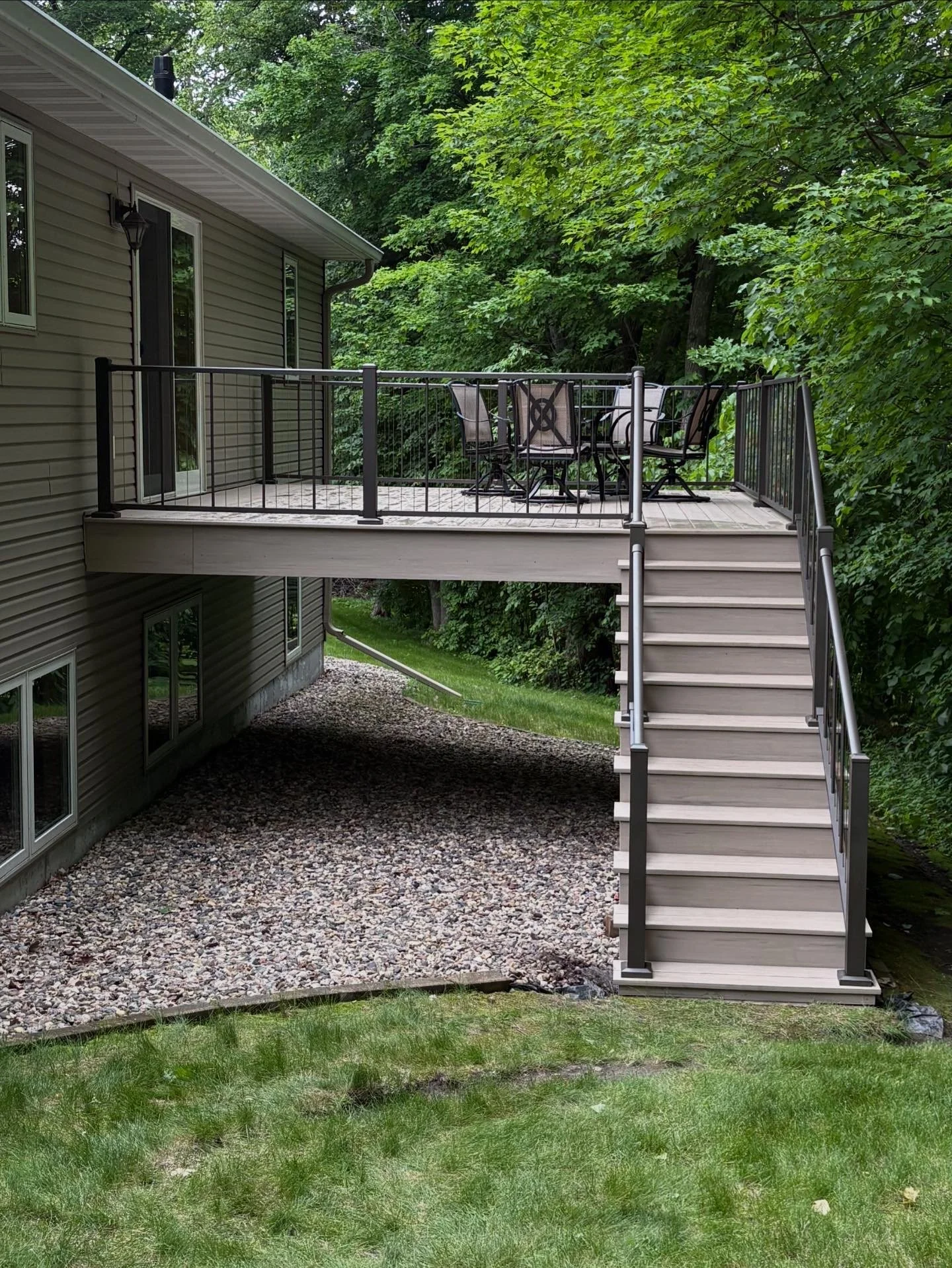 A backyard deck attached to a house, accessible by a staircase, overlooking a grassy yard and trees.
