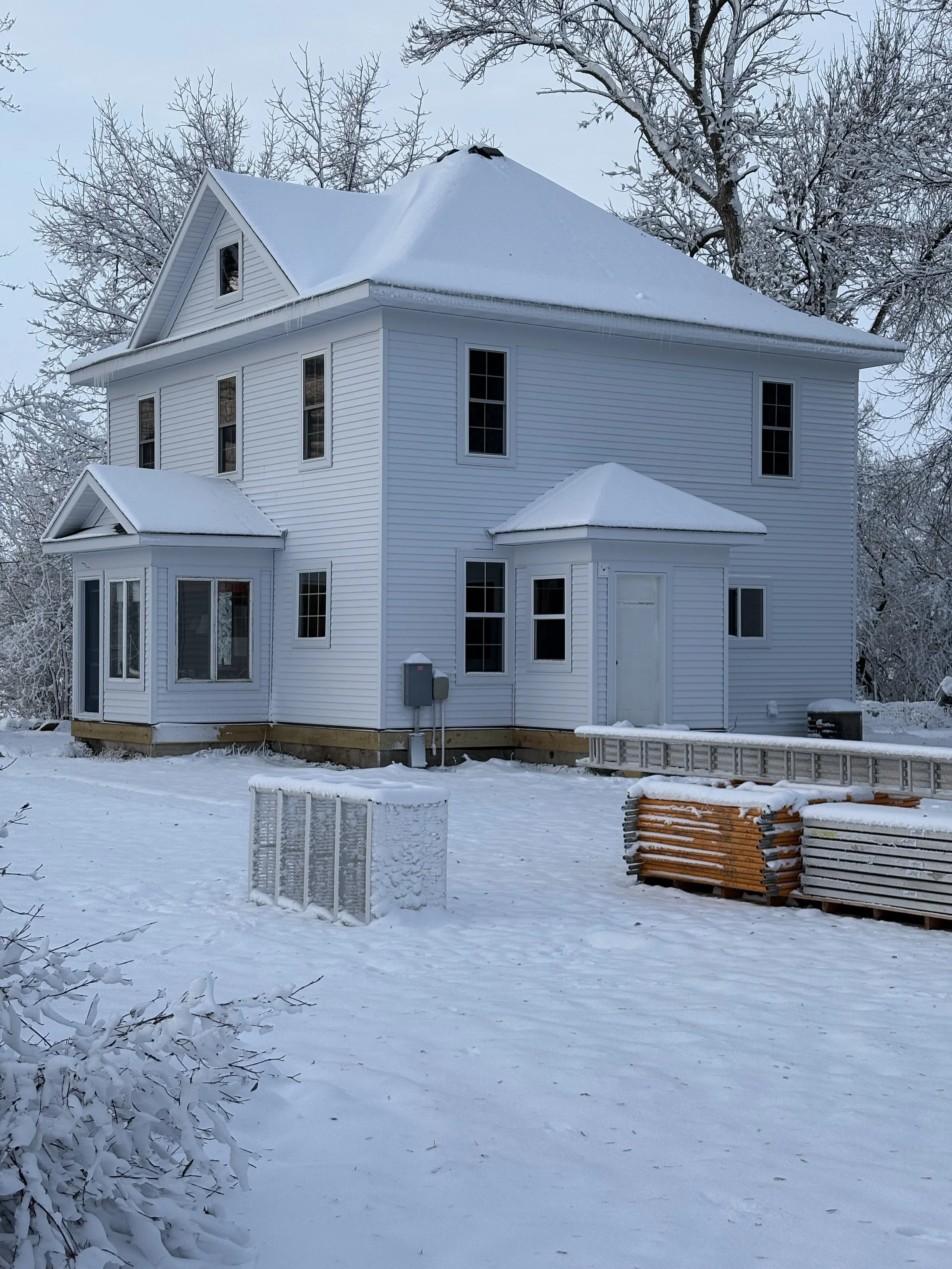 A white, two-story house with snow-covered roof and trees in the background, snow on the ground, and construction materials stacked outside.