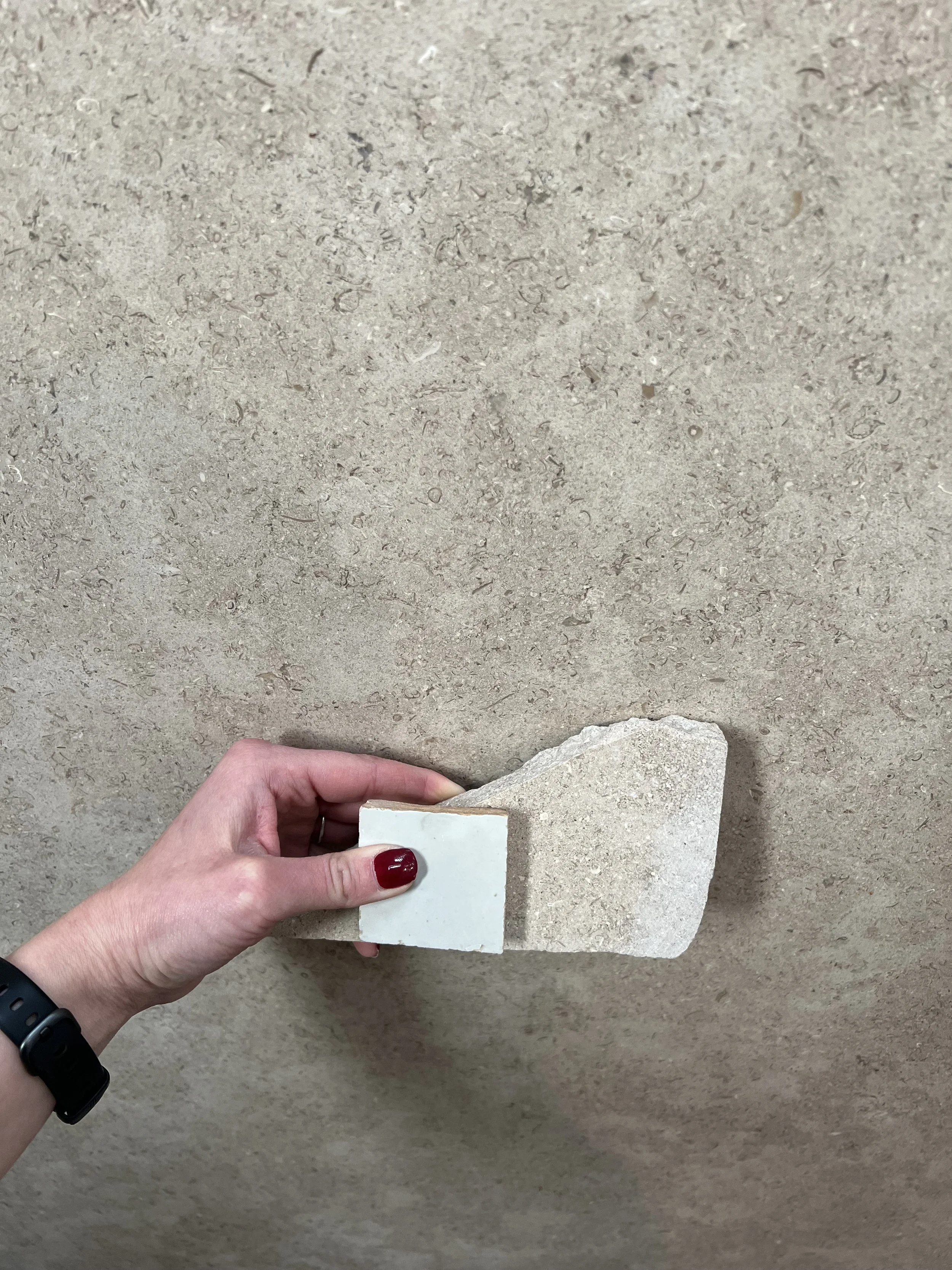 A hand holds a piece of textured stone or concrete against a similar textured wall.