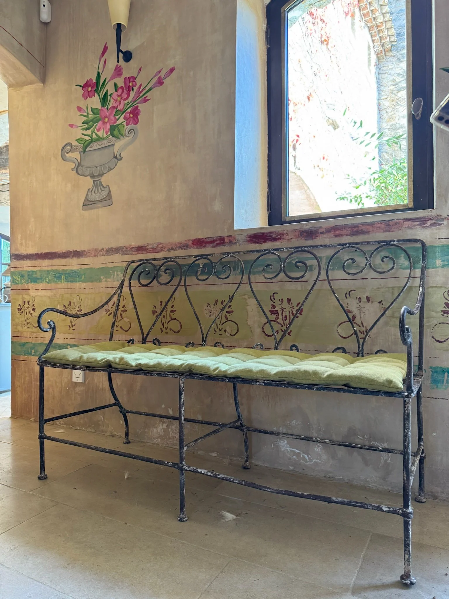 Decorative black metal bench with a yellow cushion, placed against a beige wall with painted floral art and fresco-style bands, next to a window showing an outdoor scene with a stone wall and plants.
