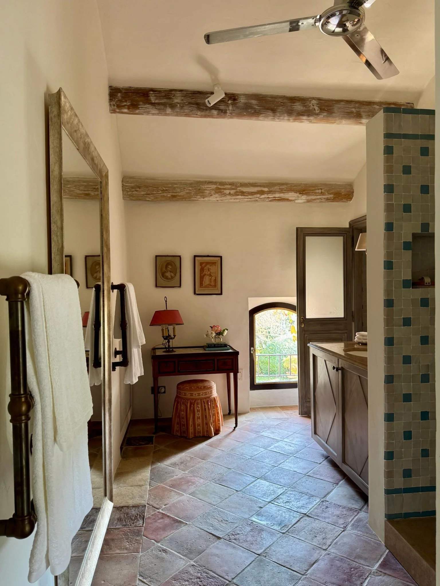 A rustic styled bathroom with stone tile flooring, a wooden ceiling with exposed beams, a ceiling fan, a mirror, towels hanging on a rack, a small wooden cabinet with a lamp and flowers, a small window with a balcony view, and a partly visible shower