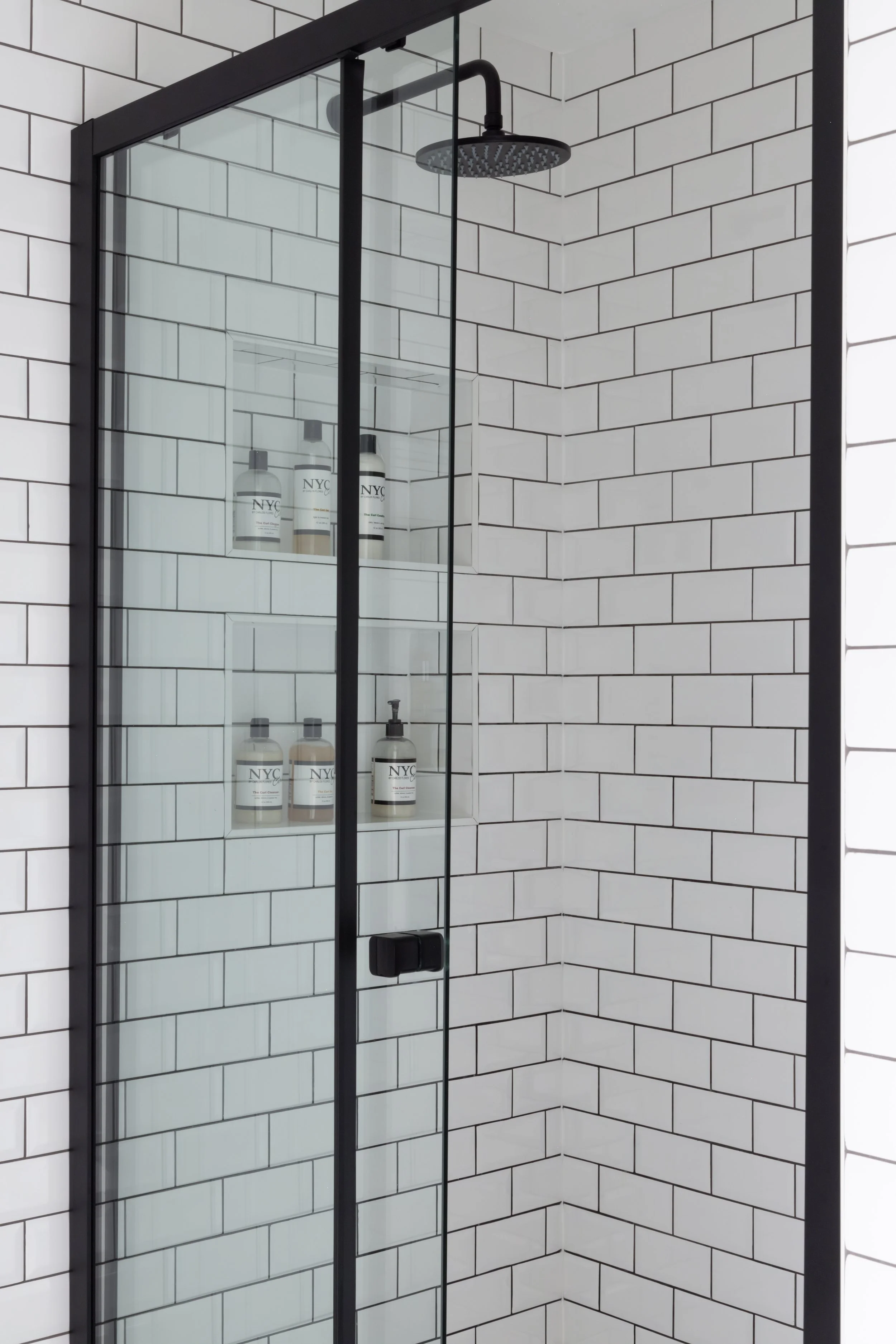 Modern shower with black frame, white subway tile walls, black showerhead, and built-in shelf with bottles of soap and shampoo.