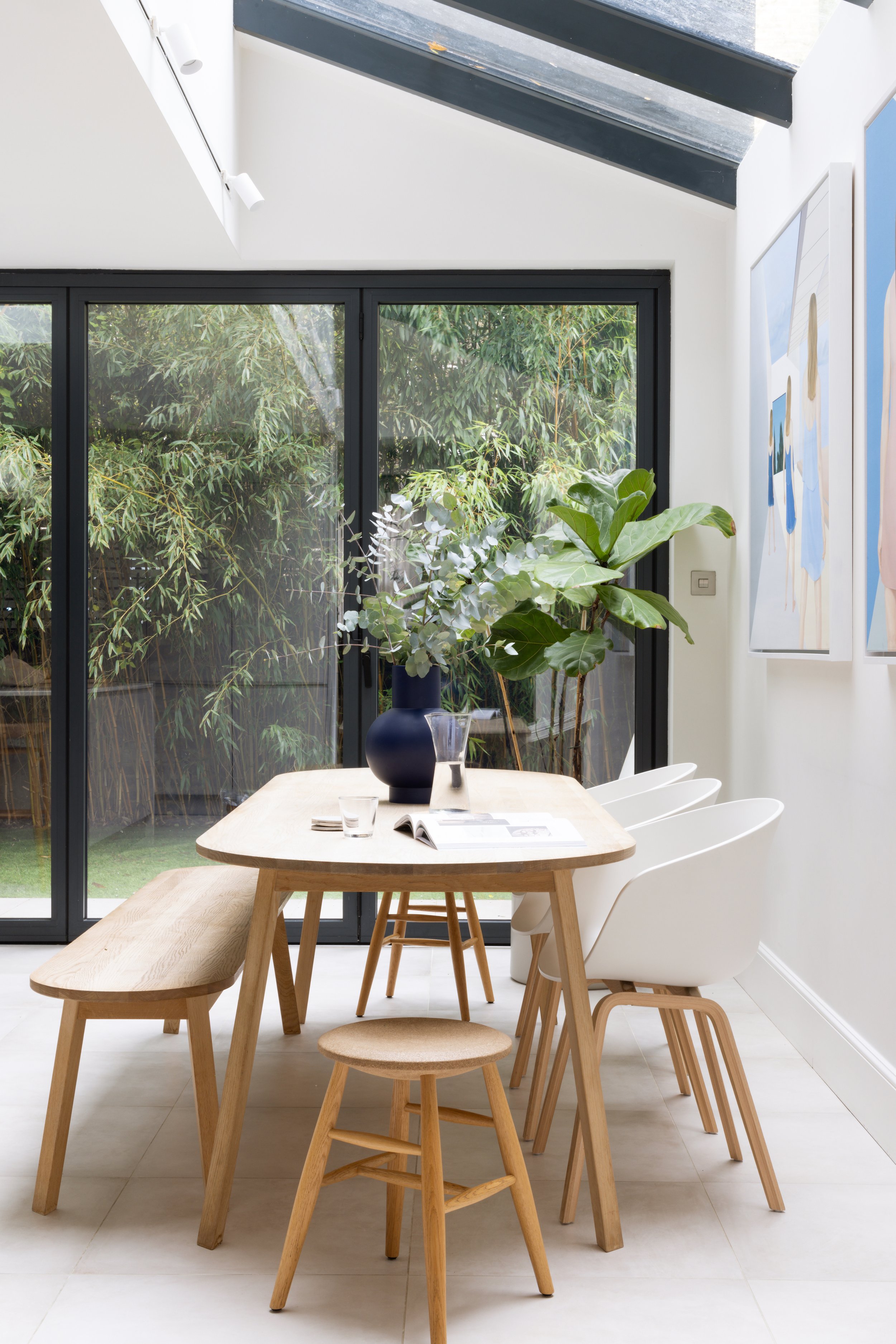Modern dining area with wooden table, white chairs, large potted plant, and glass sliding doors leading to greenery outside, with artwork on the wall.