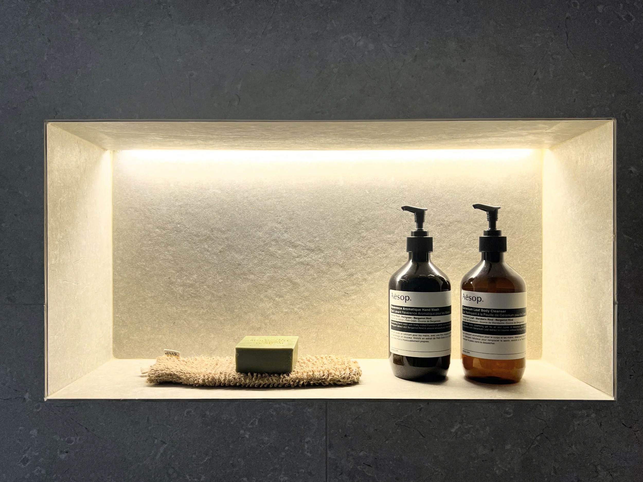 A recessed shelf with two black and amber bottles of Aesop hand wash and hand cleanser, a small beige towel with a green soap bar on top, illuminated by built-in lighting.