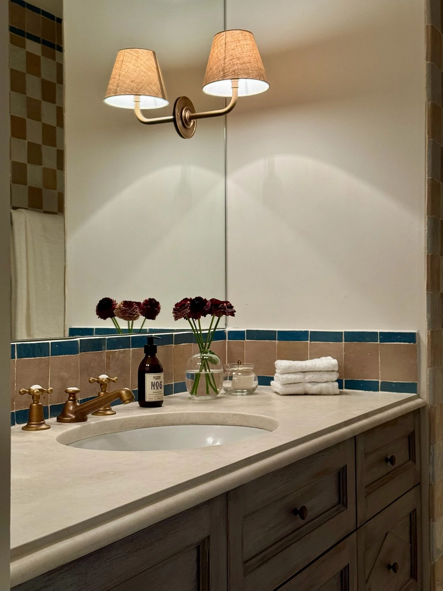 Bathroom vanity with a beige countertop, dark wood cabinet, gold faucet, a mirror, a wall-mounted twin-lamp fixture, a glass jar with dark flowers, a brown bottle, a small candle, and pulled white towels.
