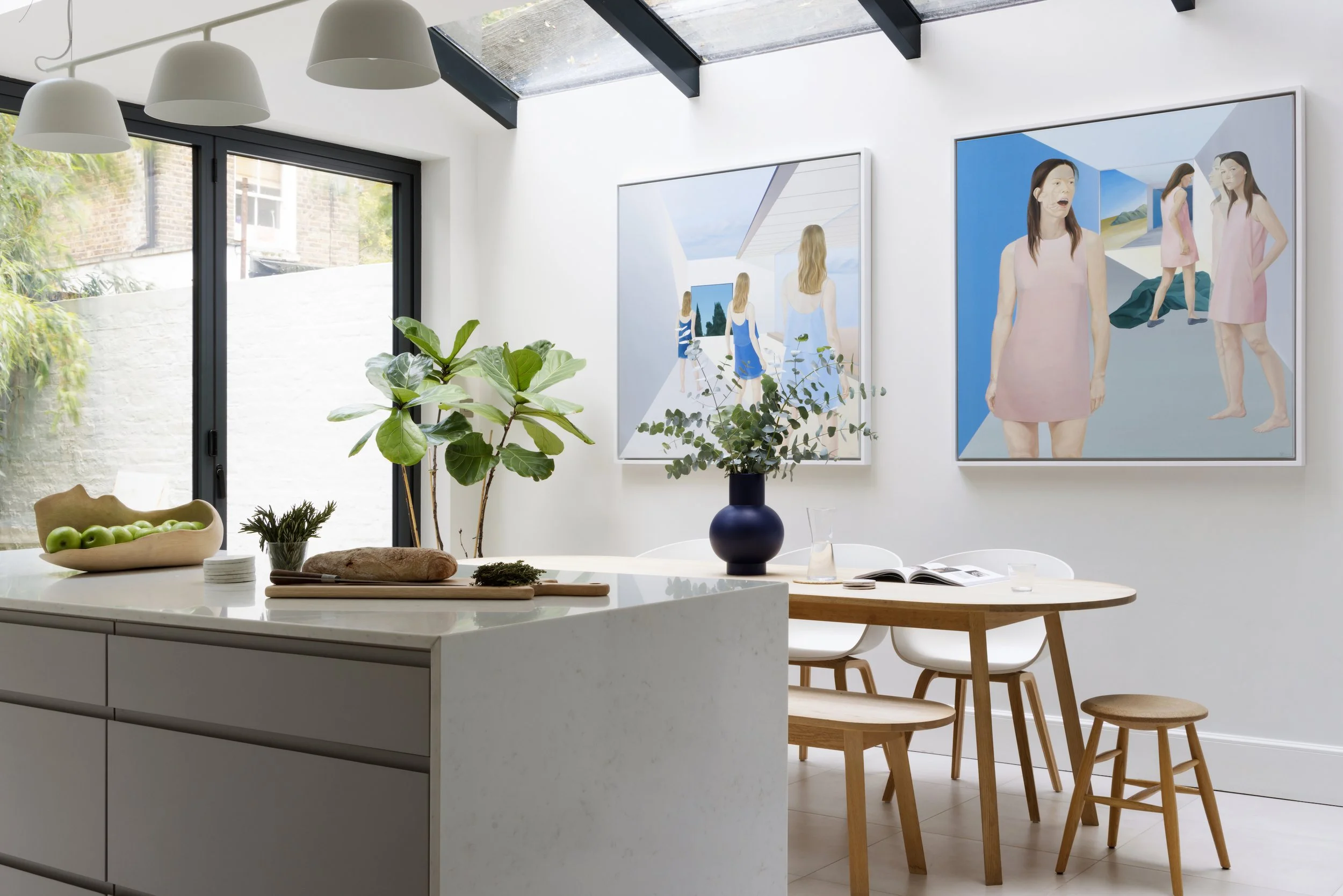 Modern kitchen and dining area with white walls, large window, green plants, wooden table with white chairs, and two large abstract art pieces on the wall.