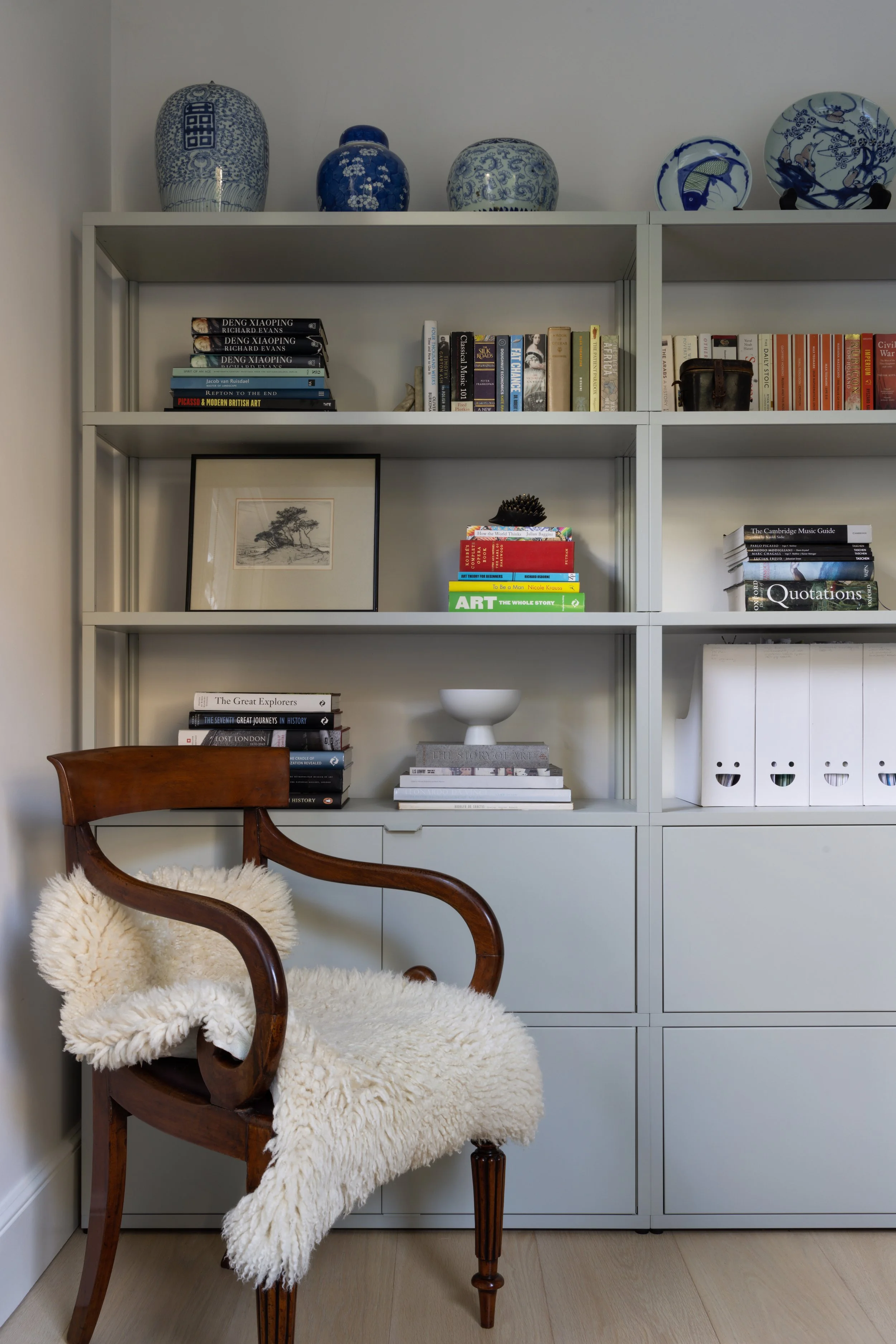 A wooden chair with a sheepskin throw in front of a light gray bookshelf filled with books, decorative plates on top, and other decorative items. Home office design, interior design, decoration, styling