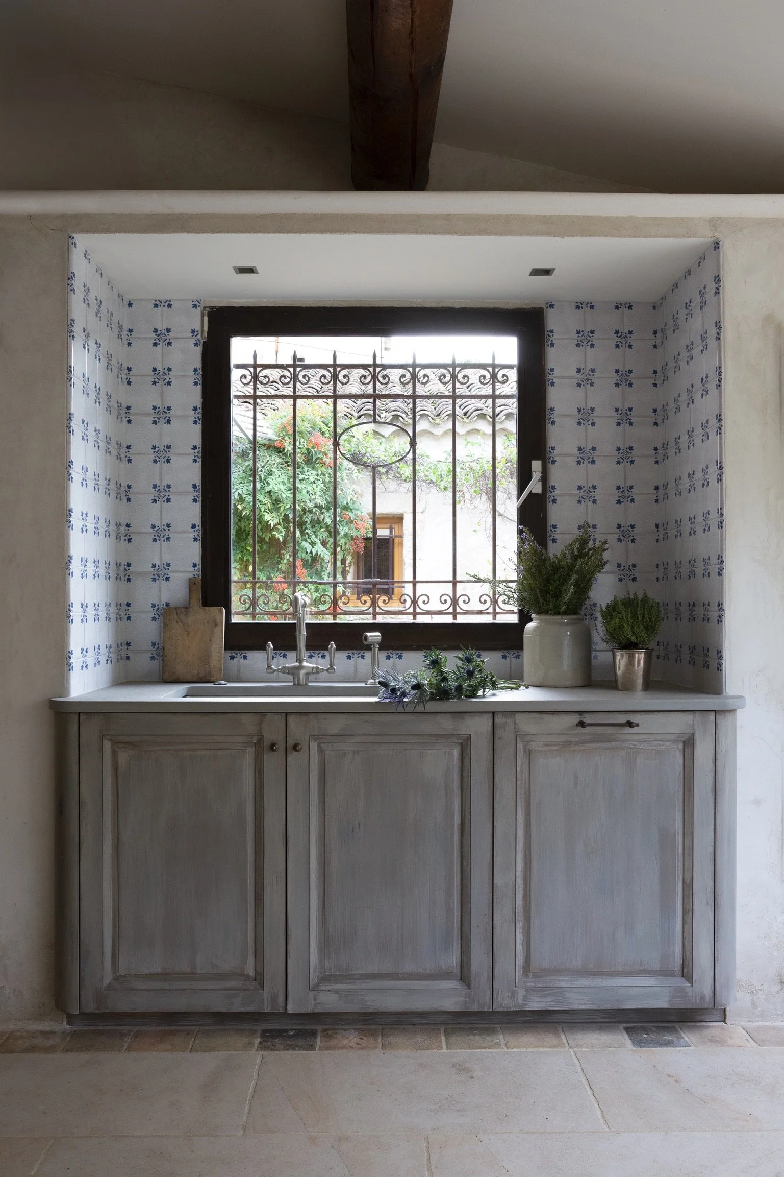 Kitchen sink area under a window with blue and white patterned tiles on the wall, decorated with potted plants.