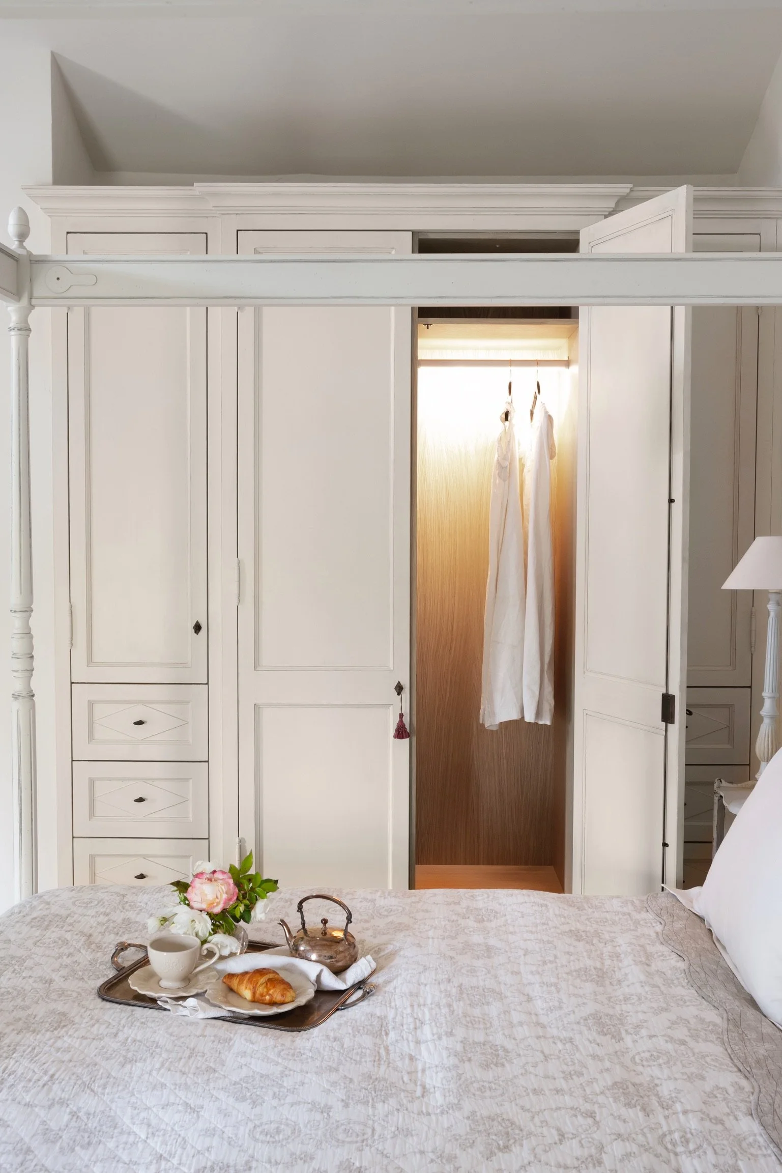 A bedroom with a white bed, a bedside lamp, and a tray with a croissant, a cup, a teapot, and flowers, overlooking a closet area with two white dresses hanging inside.