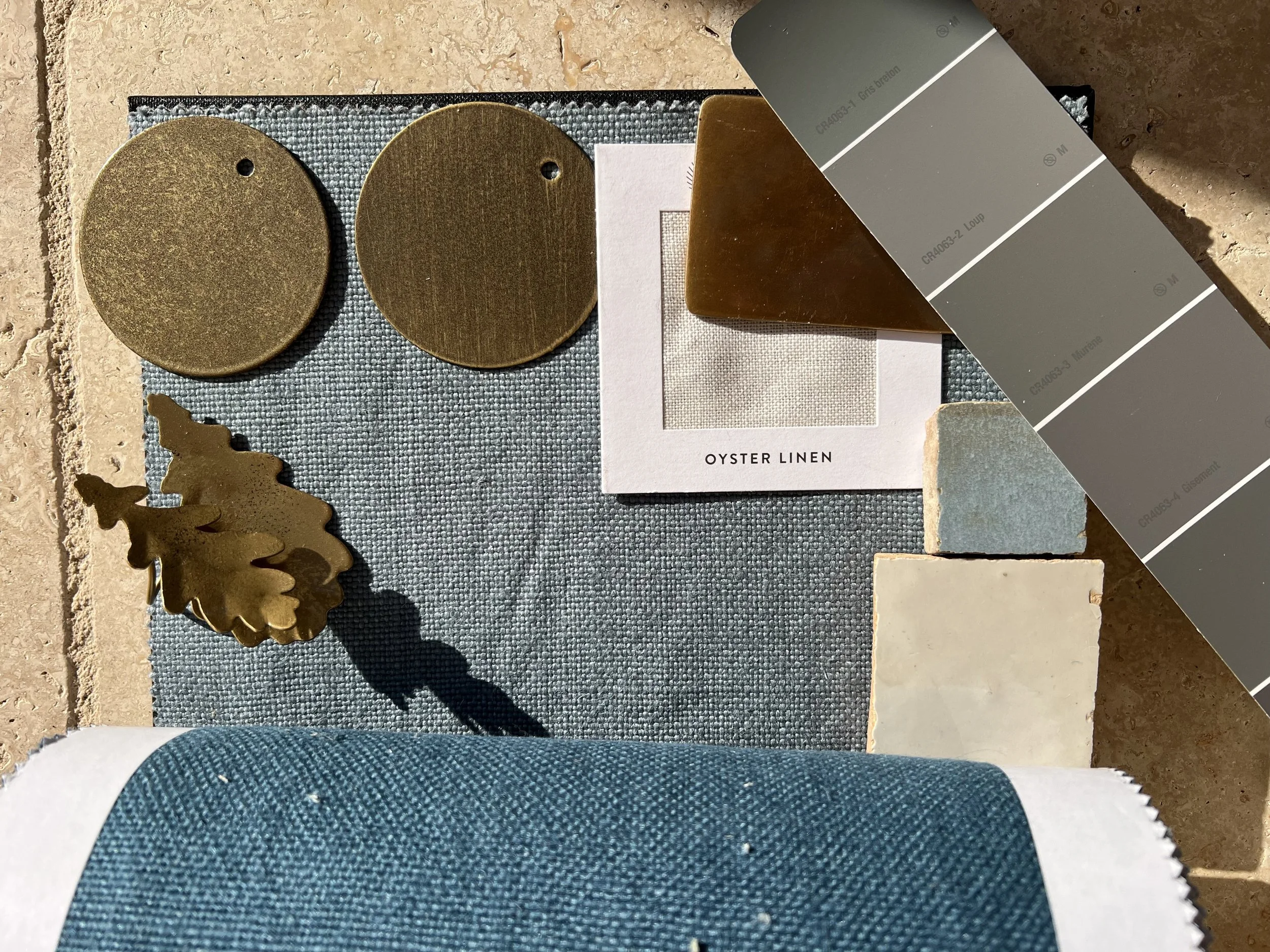 Sample materials for interior design including two round brass-colored metal pieces, a brass-colored leaf-shaped metal piece, a small fabric swatch labeled 'Oyster Linen,' a square tile with a textured surface, a larger rectangular tile, a wood sampl