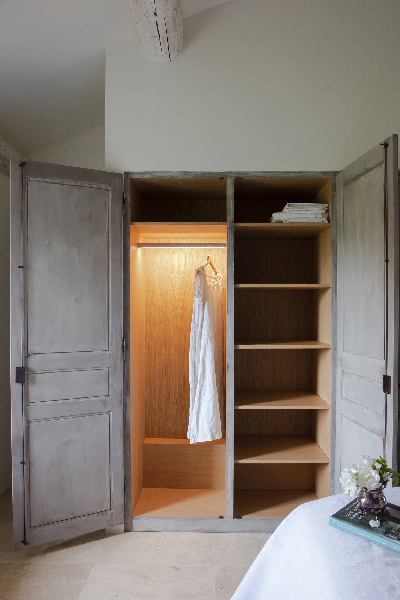 An open wooden wardrobe with distressed grey doors. The left side has a lit compartment with a hanging white dress. The right side has empty shelves with some folded clothes on the top shelf. A vase with white flowers and a book are on a nearby table