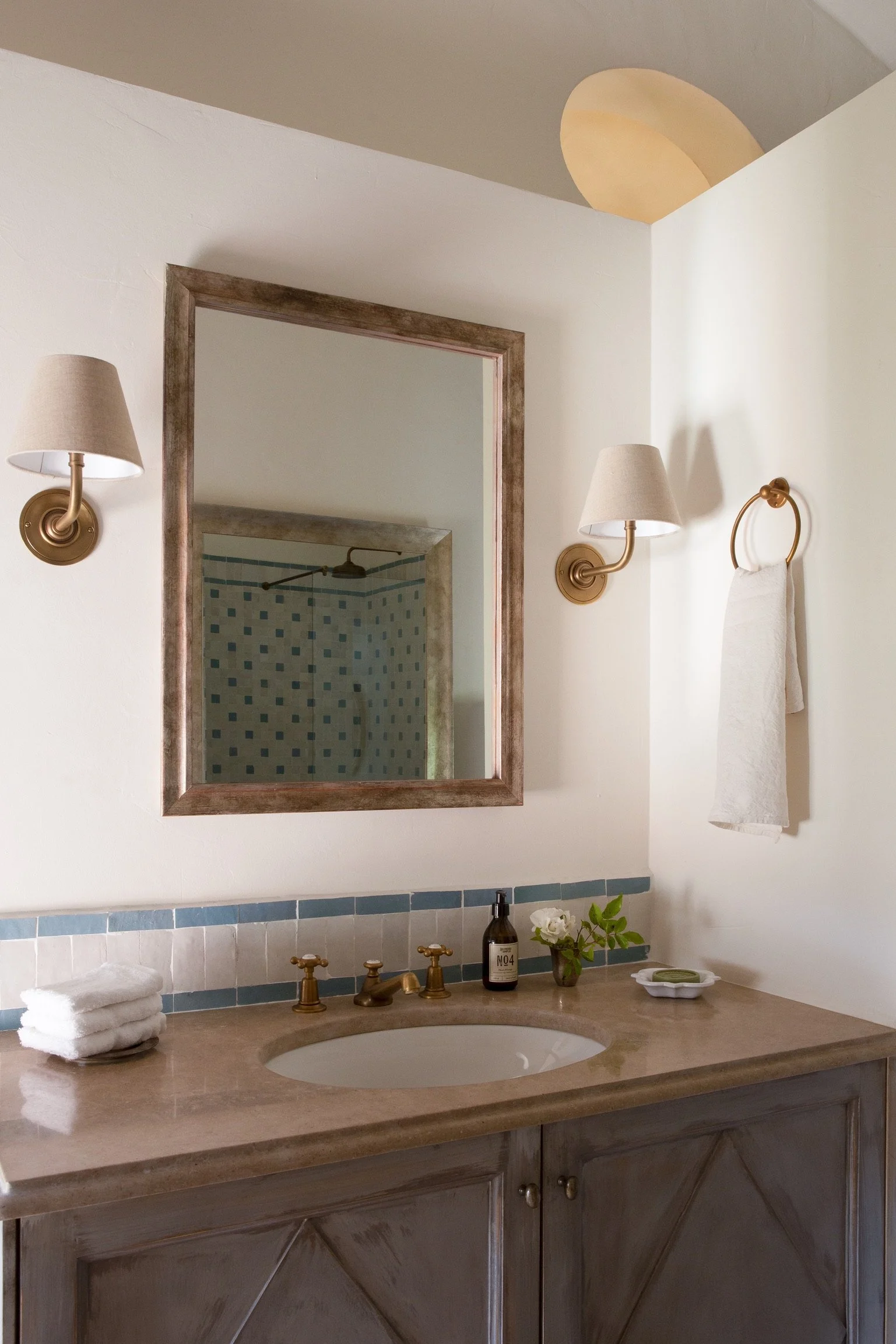 Bathroom vanity with a wooden cabinet, beige countertop, oval sink, brass faucet, framed mirror, wall-mounted lamps, towel ring with a towel, soap dish, soap bottle, and small flower arrangement.