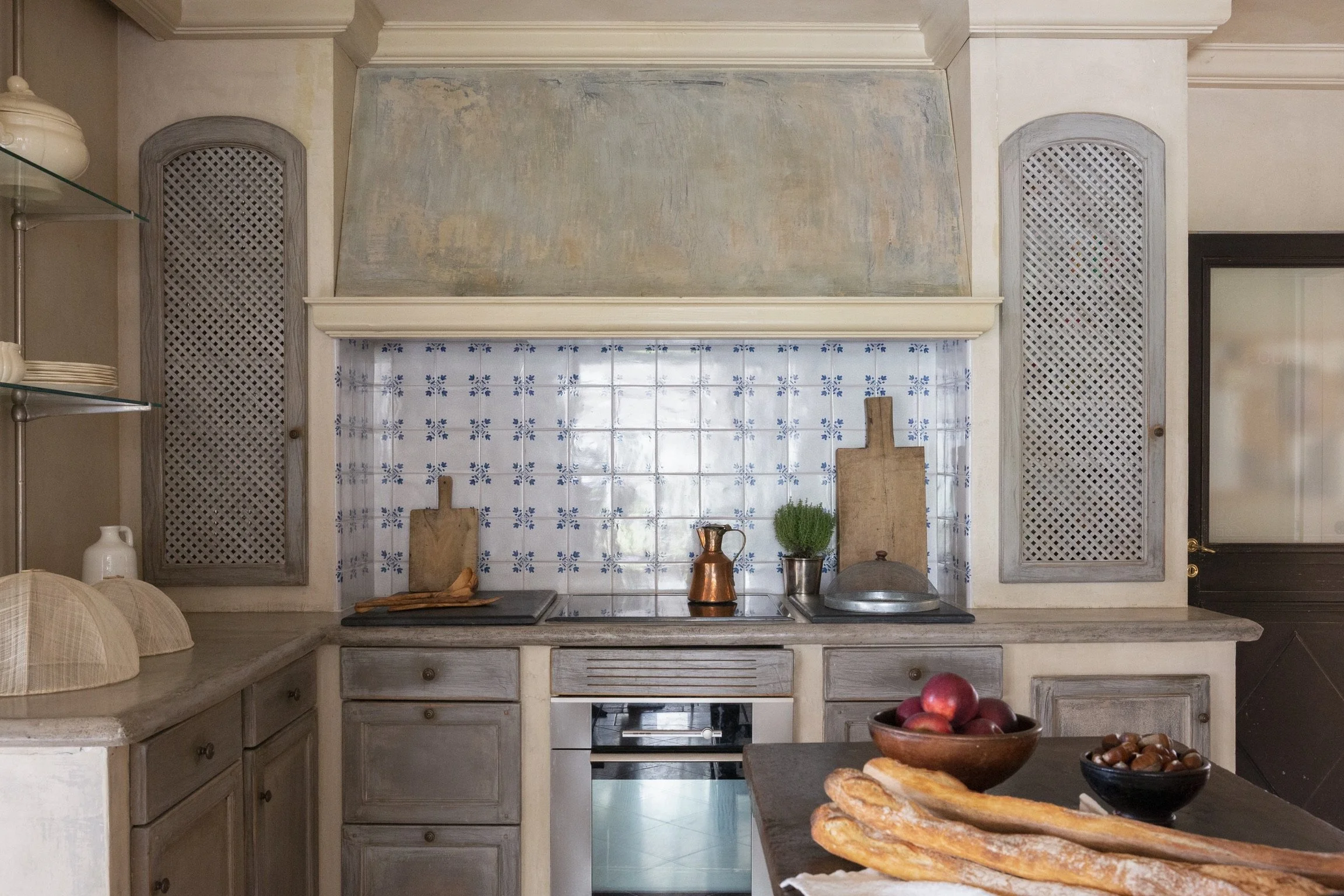 A rustic kitchen with a tiled backsplash, wooden cabinets, and a stove, decorated with cutting boards, a copper pitcher, herbs, and bowls of fruit and bread.