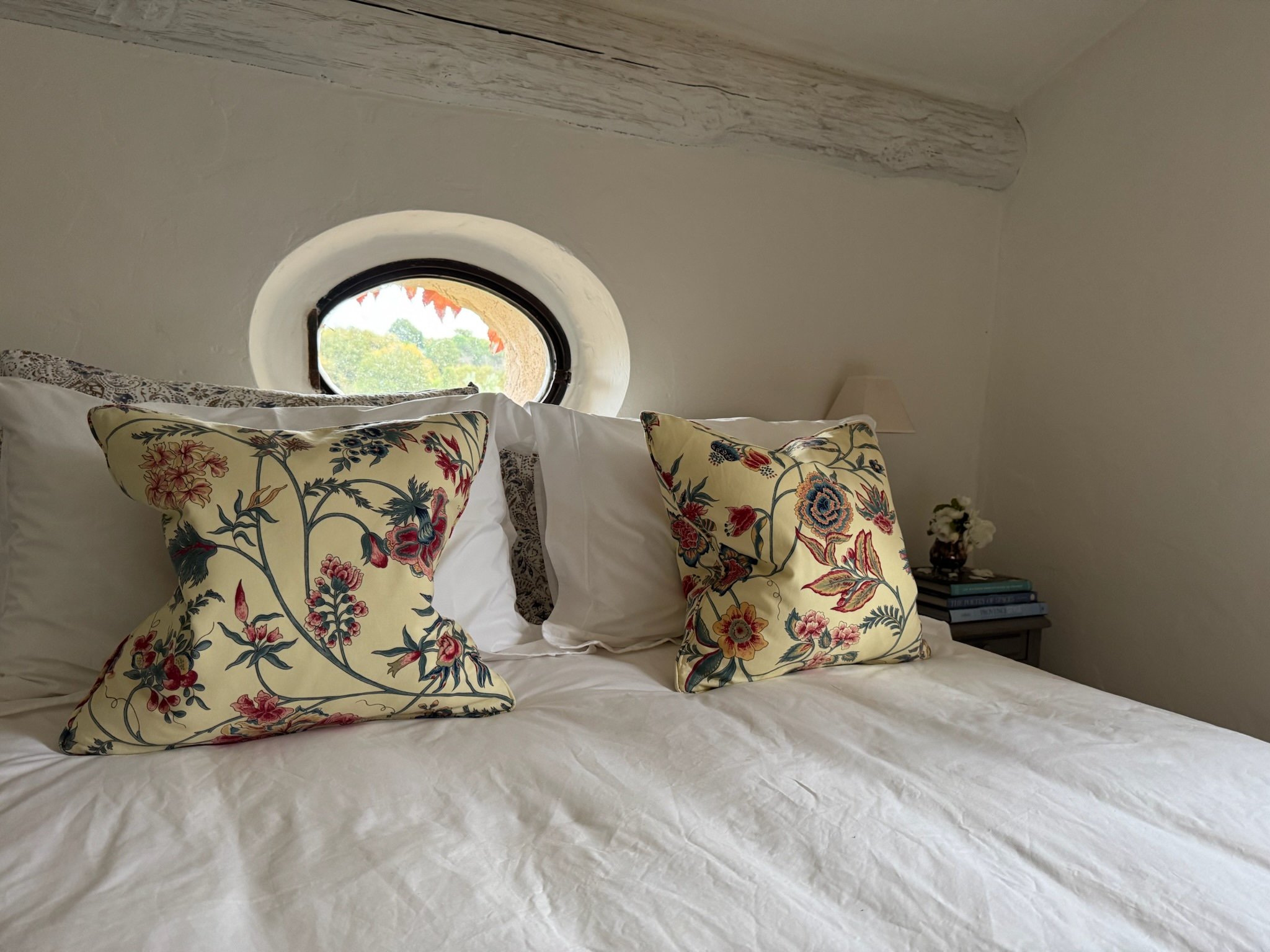 A bed with white linens and two floral pillows, in a room with a small circular window and a bedside table with books and flowers.