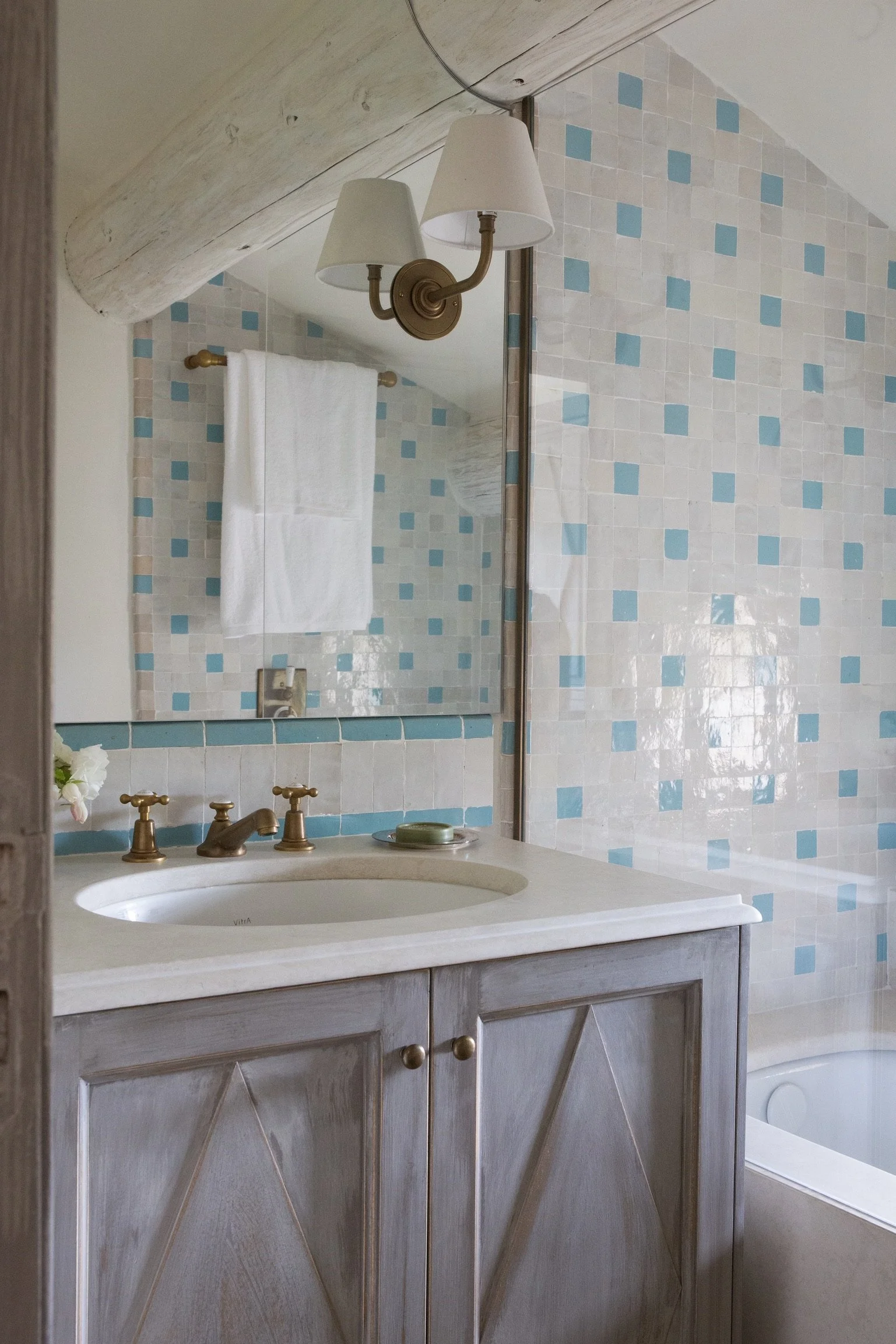 A bathroom with a vanity, countertop, mirror, and a wall with beige and blue mosaic tiles. There is a brass wall sconce with two lampshades above the mirror and a white towel hanging on a brass rod.