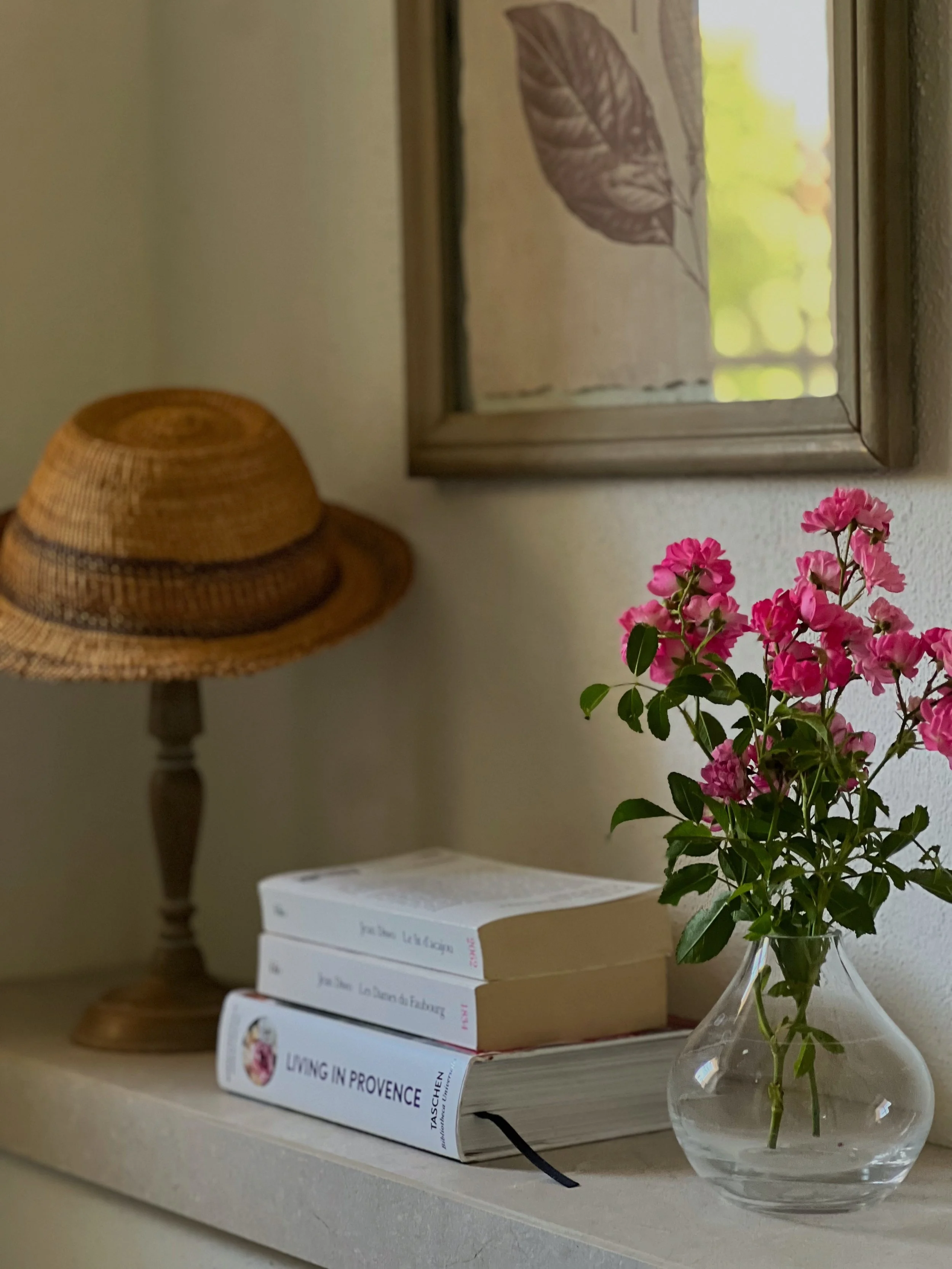 A close-up of a beige shelf holding a stack of three books, a transparent glass vase with pink flowers, a woven rattan hat on a wooden lamp base, and a window with sheer curtains featuring leaf patterns in the background.