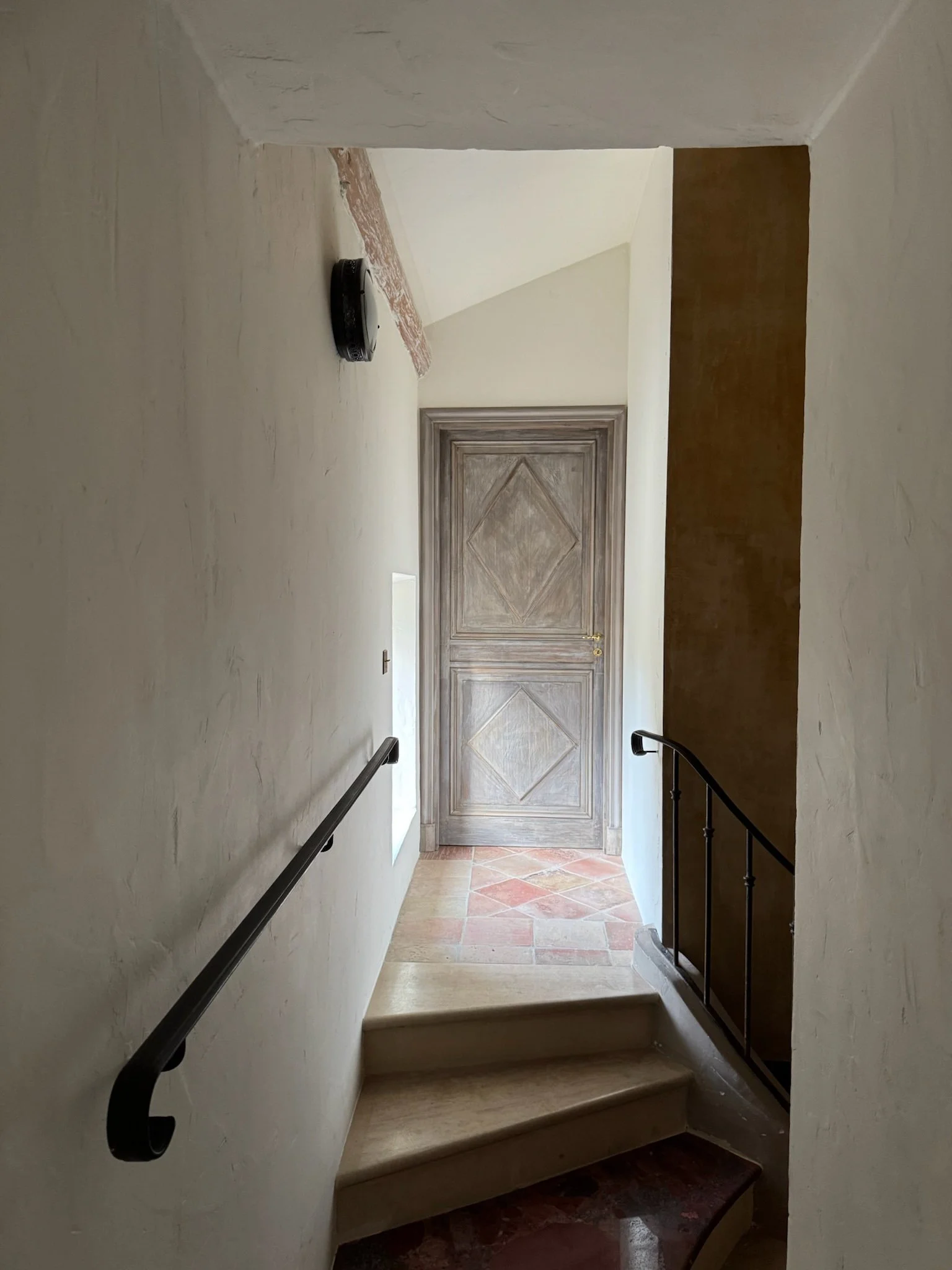 Interior view of a staircase leading to a wooden door at the top, with beige walls, a black handrail on the left, a small window on the left wall, and a terracotta tiled floor.