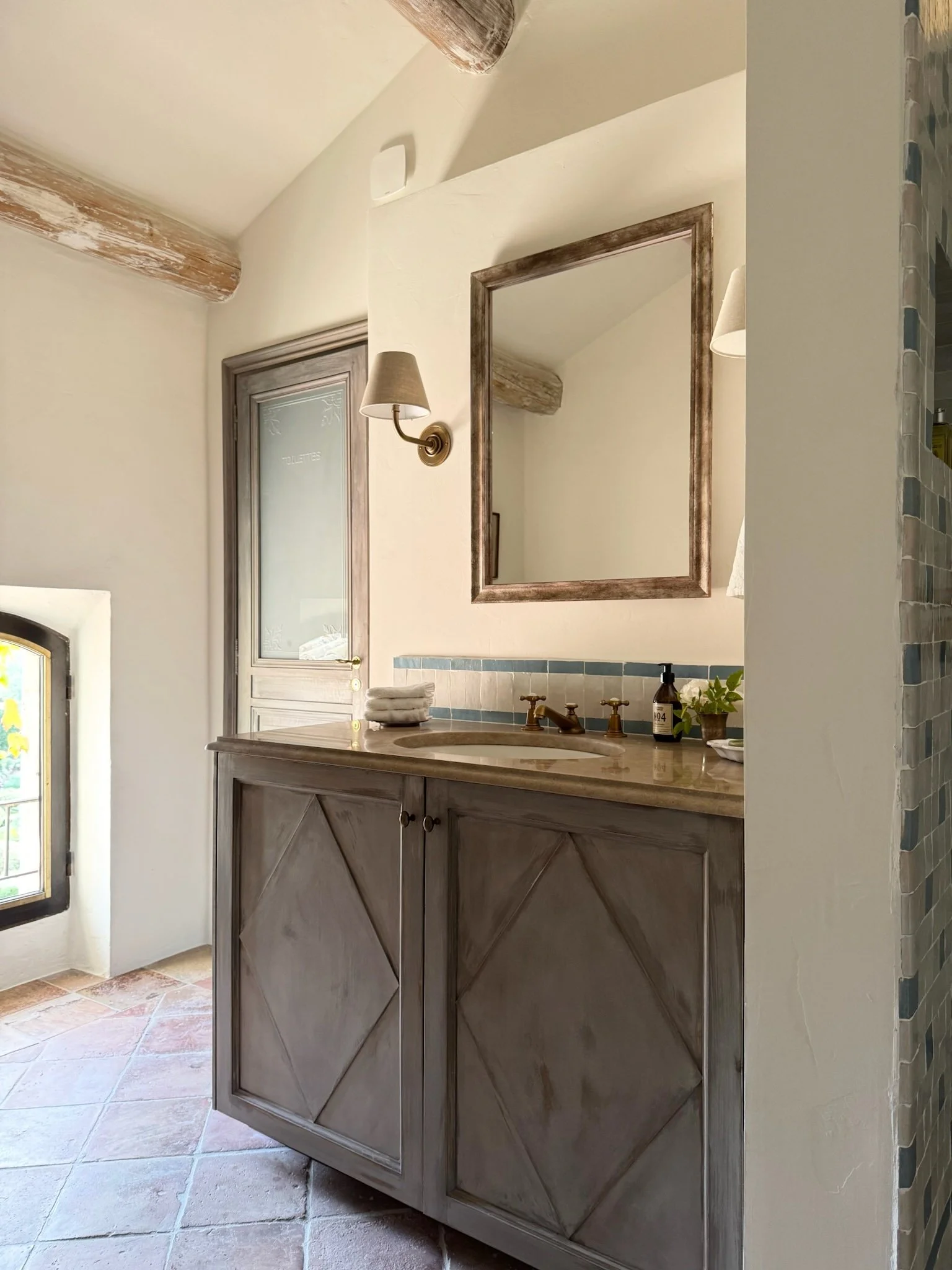 Bathroom vanity with a framed mirror, wall sconces, and a decorated countertop, near a frosted glass door and a small window with black trim, in a rustic style.