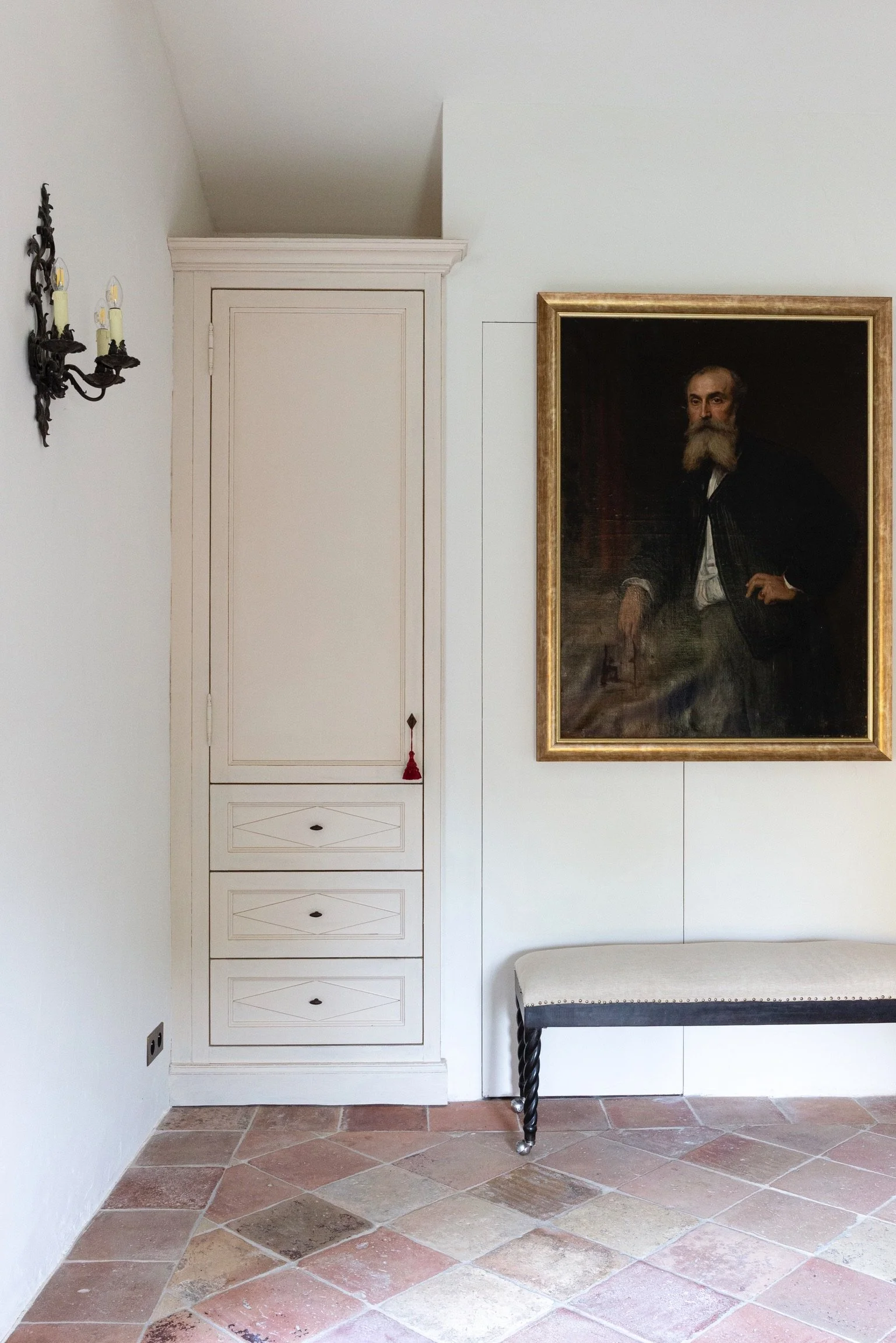 A white wall with a black wall sconce holding three candles on the left, a tall white cabinet with three drawers and a decorative red tassel hanging from the top door, a large framed portrait of a bearded man on the right, and a black and white uphol
