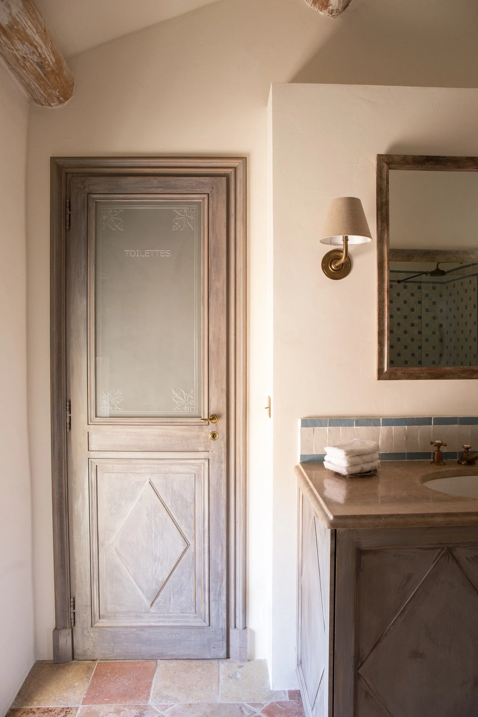 A wooden door with frosted glass panel marked 'Toilettes,' next to a bathroom vanity with folded towels, a mirror, and a wall-mounted light fixture.