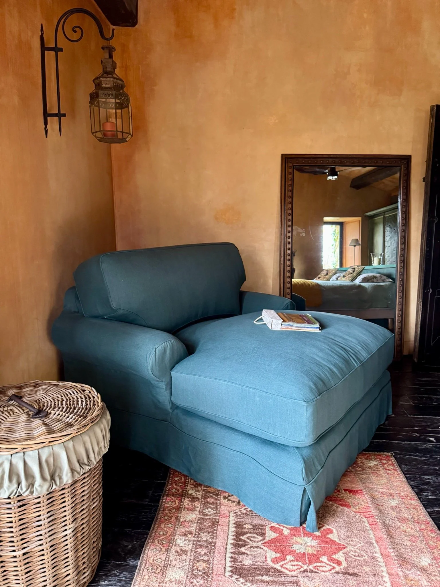 A teal lounge chair with a slipcover placed on a patterned area rug in a cozy room with warm-colored painted walls, a wicker laundry basket, a large mirror reflecting part of the room, and a wall-mounted lantern-style light fixture.