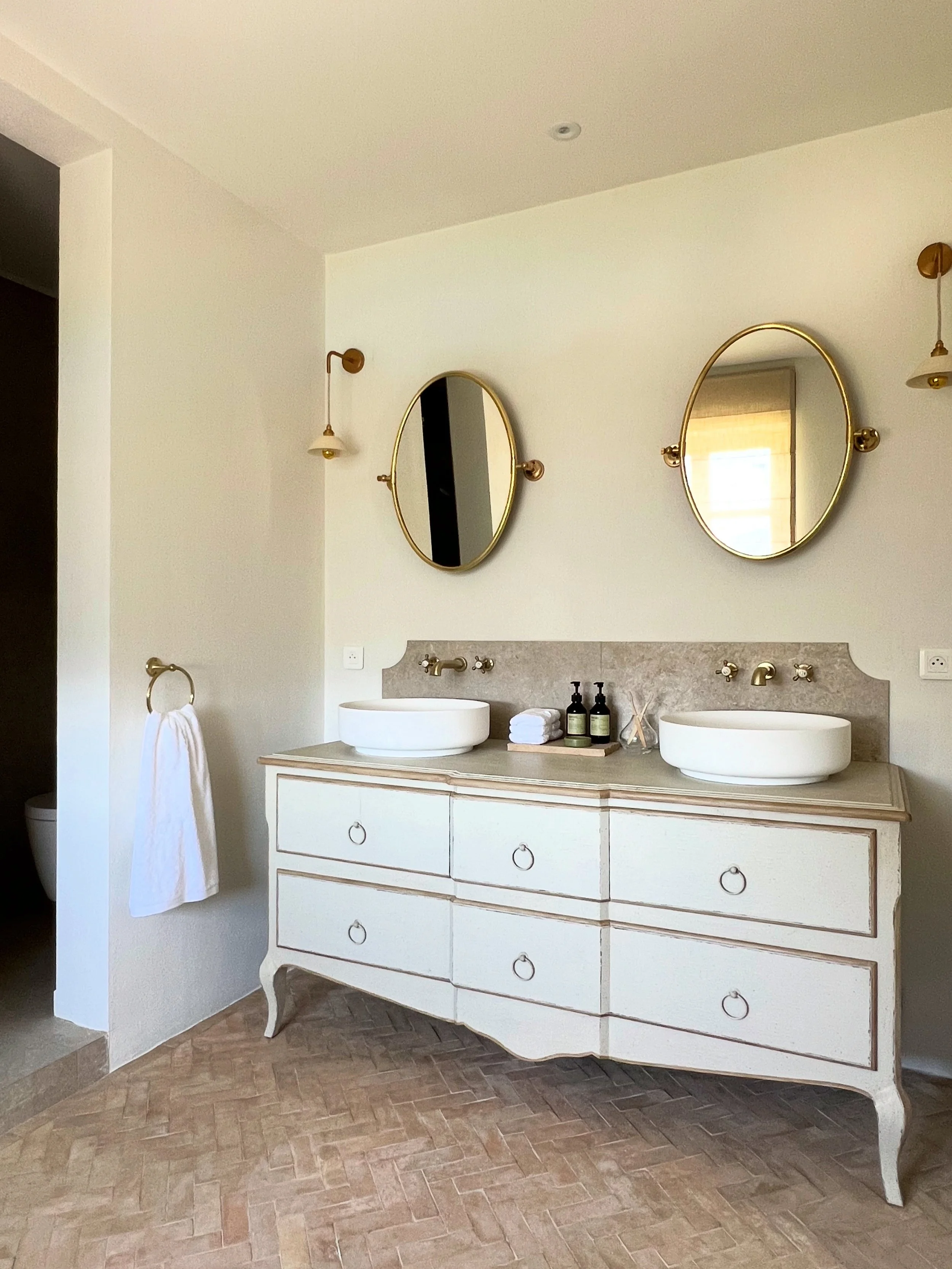 A bathroom vanity with two round vessel sinks, two oval mirrors with  frames, and a beige piece of marble behind the sinks. There are towels, soap, lotion, and reed diffusers on the counter, with a white towel hanging on a gold towel ring. The floor 