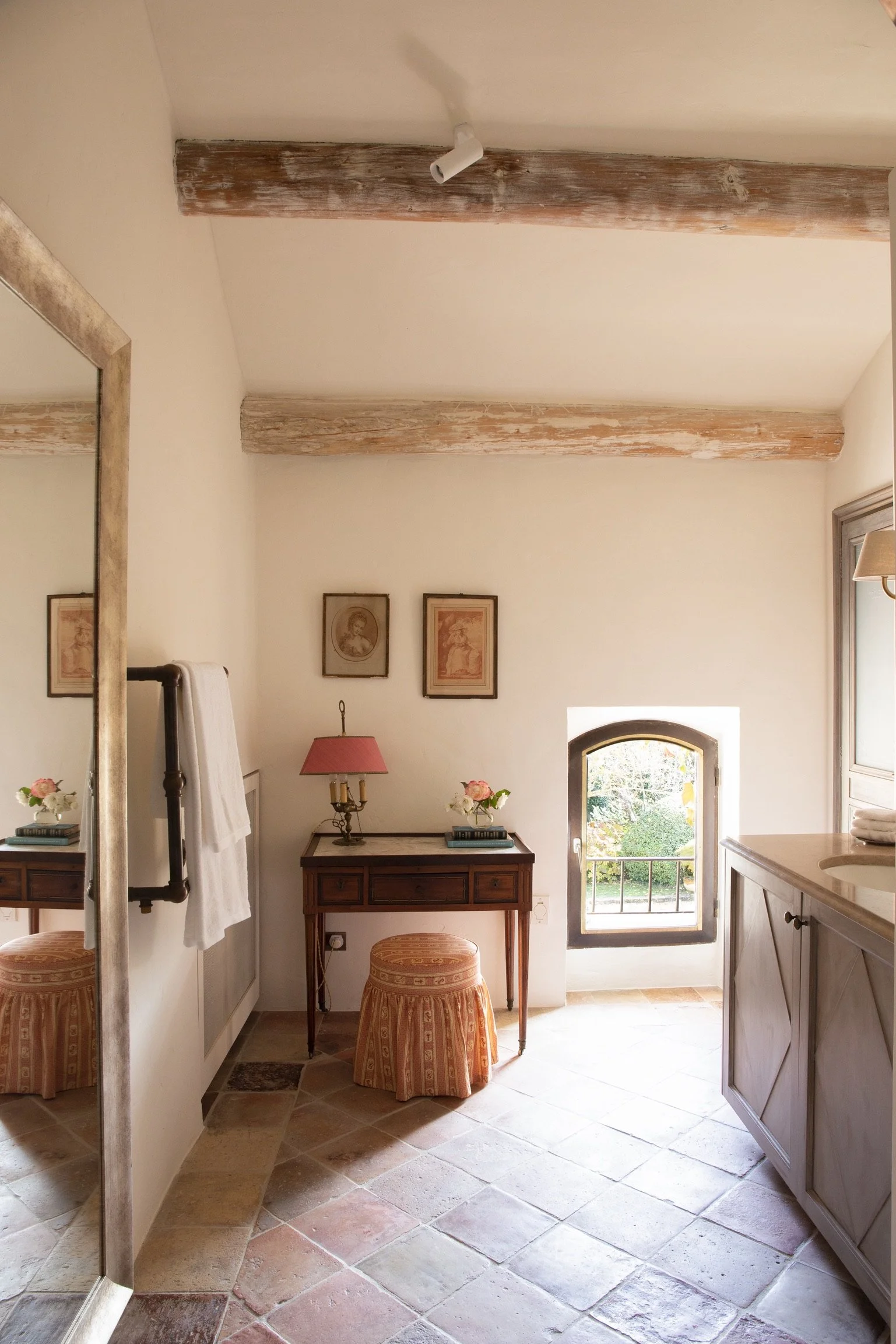 A cozy corner of a room with a small window, a wooden table with pink flower arrangements, framed artwork on the wall, and rustic ceiling beams, with a mirror reflecting part of the room on the left.