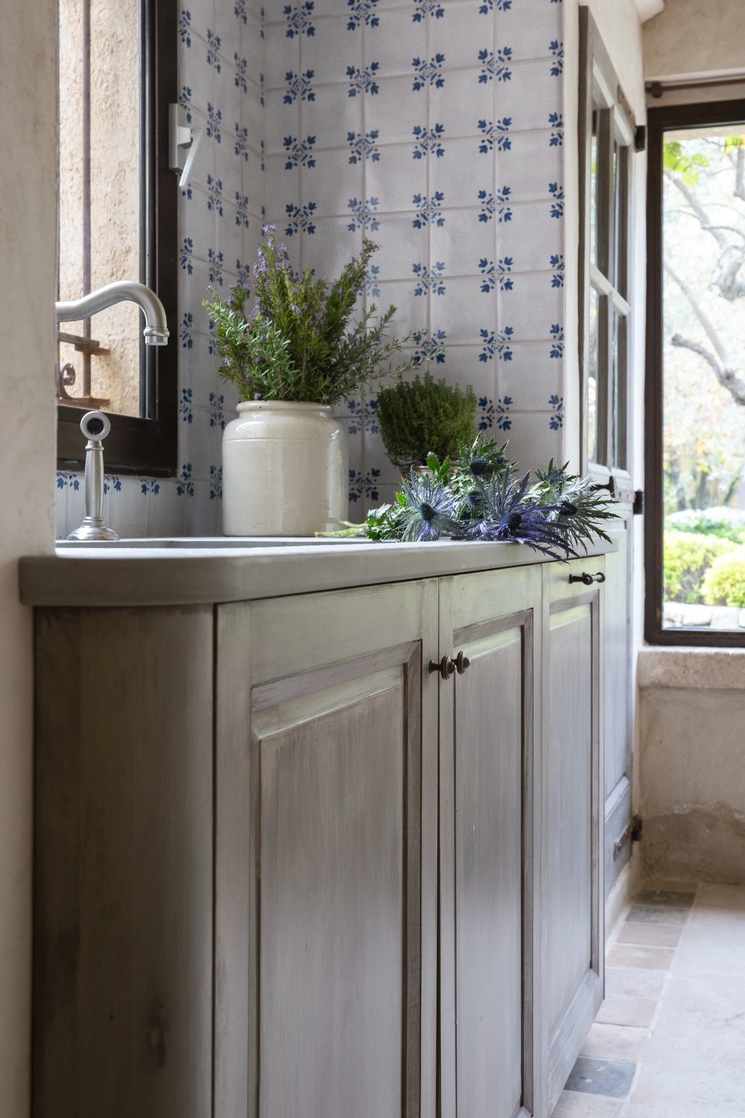 A rustic kitchen or laundry area with a stone countertop, light wood cabinet doors, and a white ceramic sink. A plant with purple flowers and green foliage sits in a cream-colored jar on the counter. Blue and white patterned tiles decorate the wall b