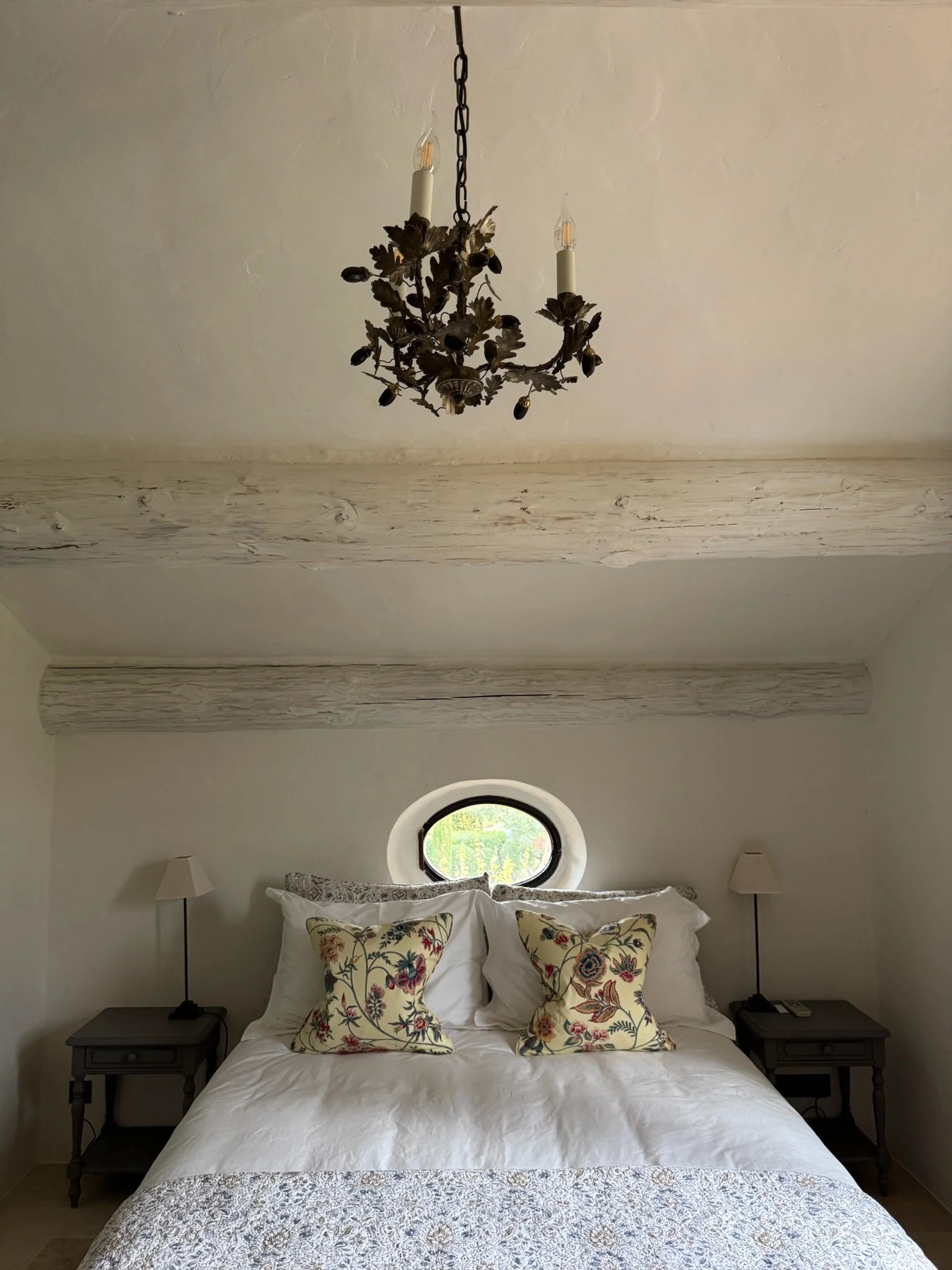 A bedroom with a white bed, decorative pillows, two black nightstands with lamps, a small oval window, and a chandelier hanging from the ceiling.