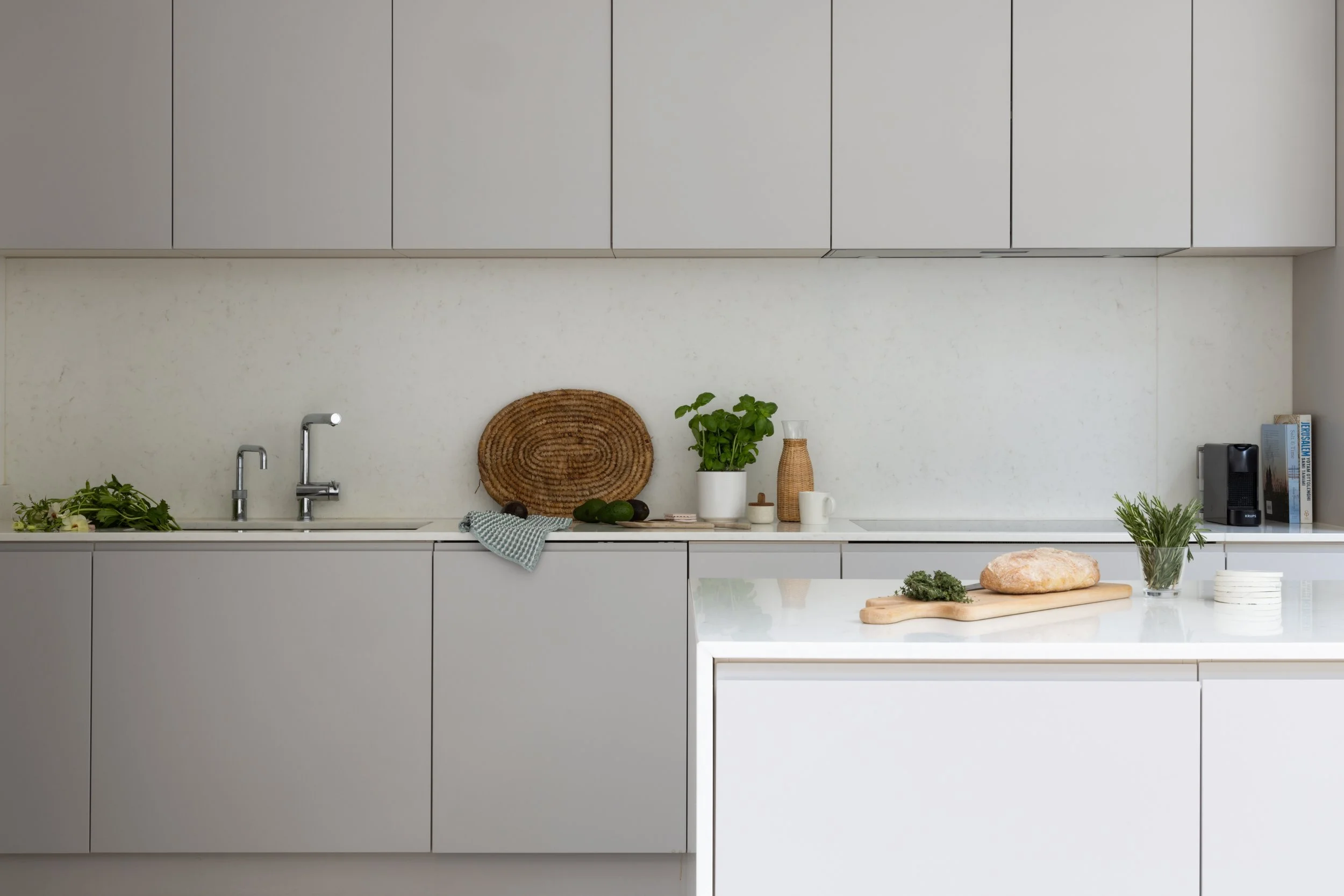 kitchen designer, Minimalist white kitchen with countertops holding a bread loaf, herbs, potted plants, and a cutting board, with white cabinetry above.
