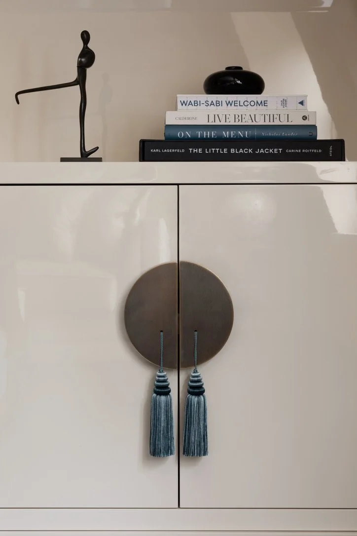 Close-up of a white cabinet with two turquoise tassels hanging from round metallic door pulls, and a decorative sculpture, books, and a vase on top.