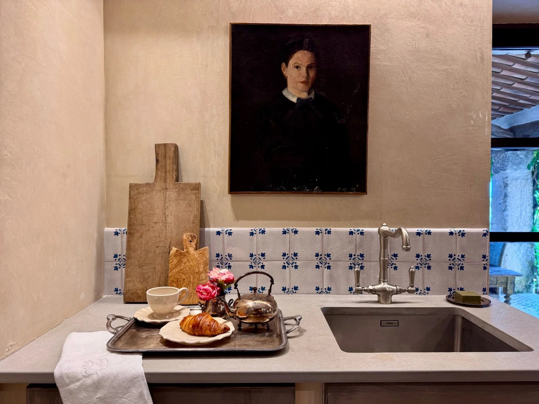 A cozy kitchen countertop with a tray holding a croissant, a cup, a small flower vase, and a teapot. Behind the tray are wooden cutting boards leaning against a beige wall. The backsplash features white tiles with blue floral patterns, and above it h