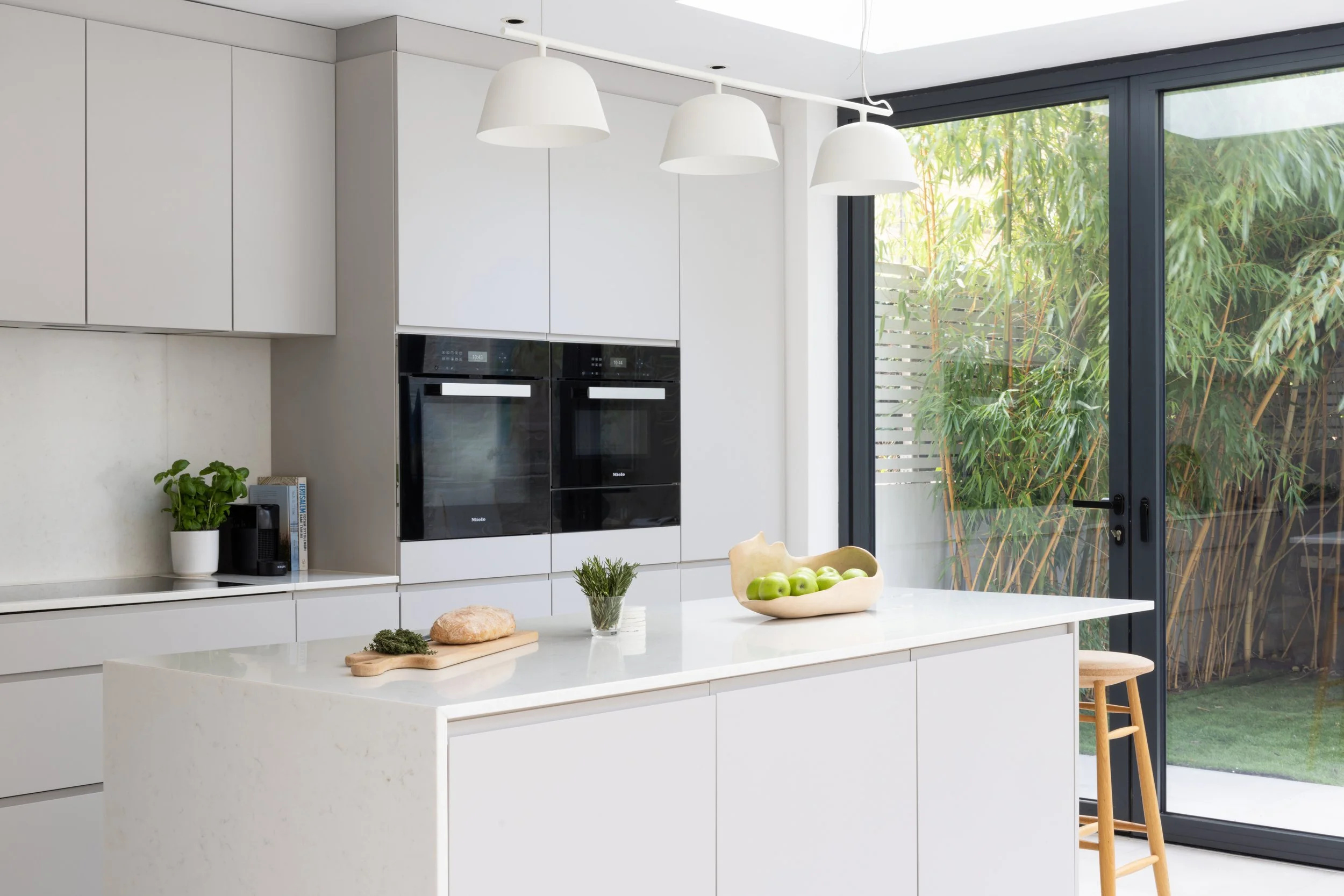 Modern white kitchen with an island, black double ovens, large sliding glass door leading to a garden, hanging white pendant lights, green apples in a bowl, potted plant, and wooden bar stool.