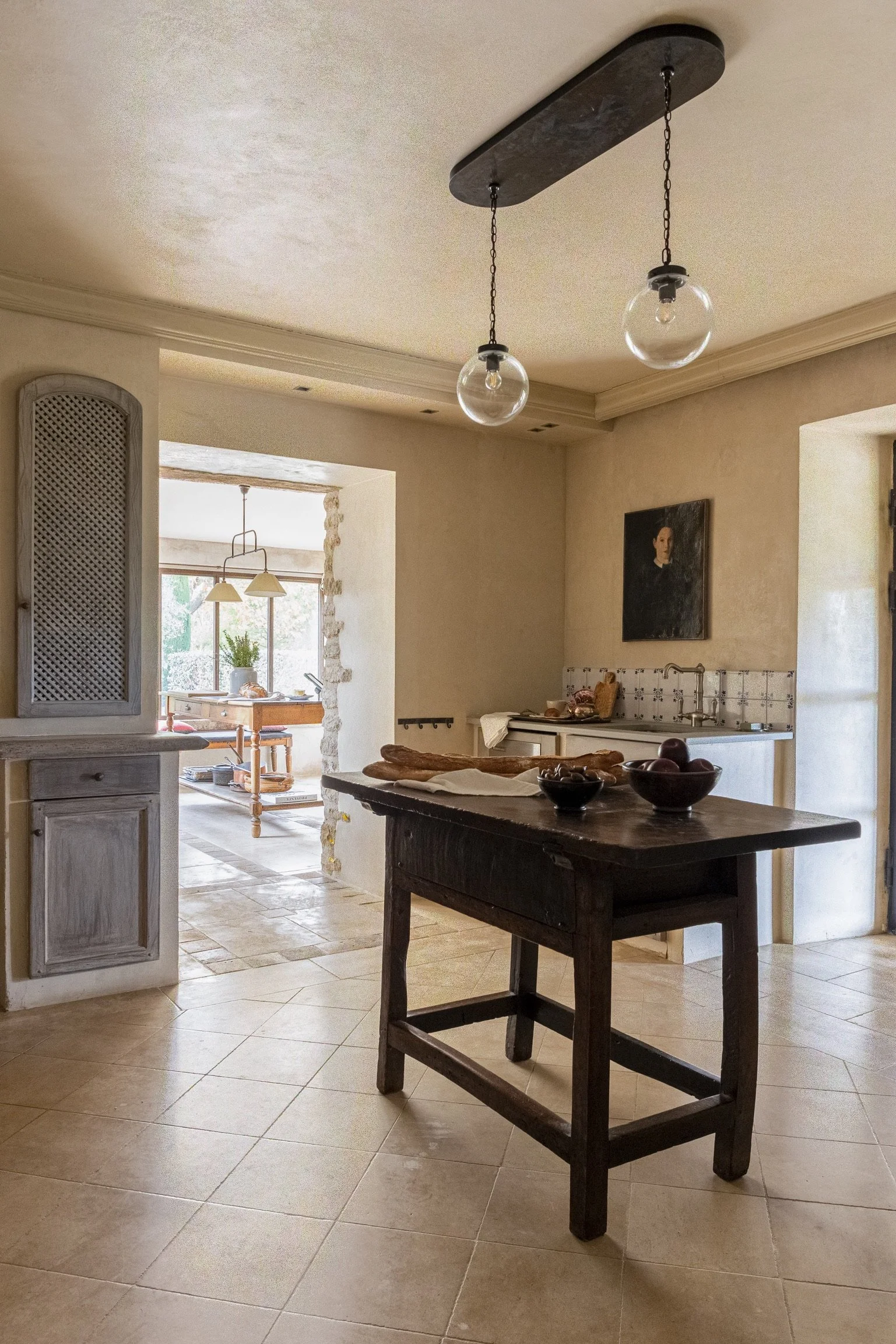 View of a cozy kitchen with beige walls and tiled flooring, featuring a black wooden kitchen island, hanging pendant lights, and a small wall-mounted sink with a tile backsplash. In the background, a dining area with a table and chairs is visible thr