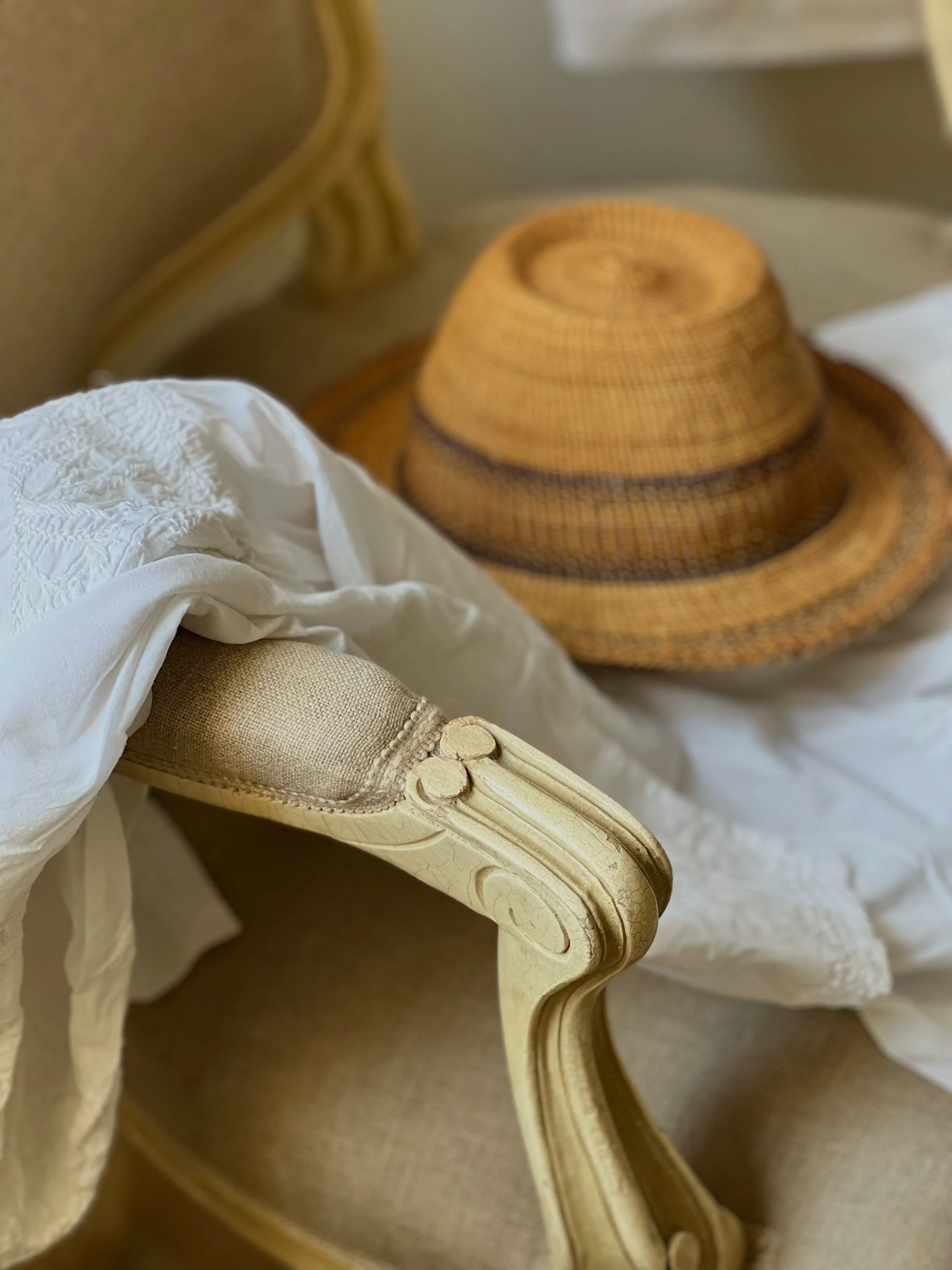 A beige vintage armchair with an ornate wooden armrest, white fabric draped over it, and a straw hat with a wide brim resting on the fabric.