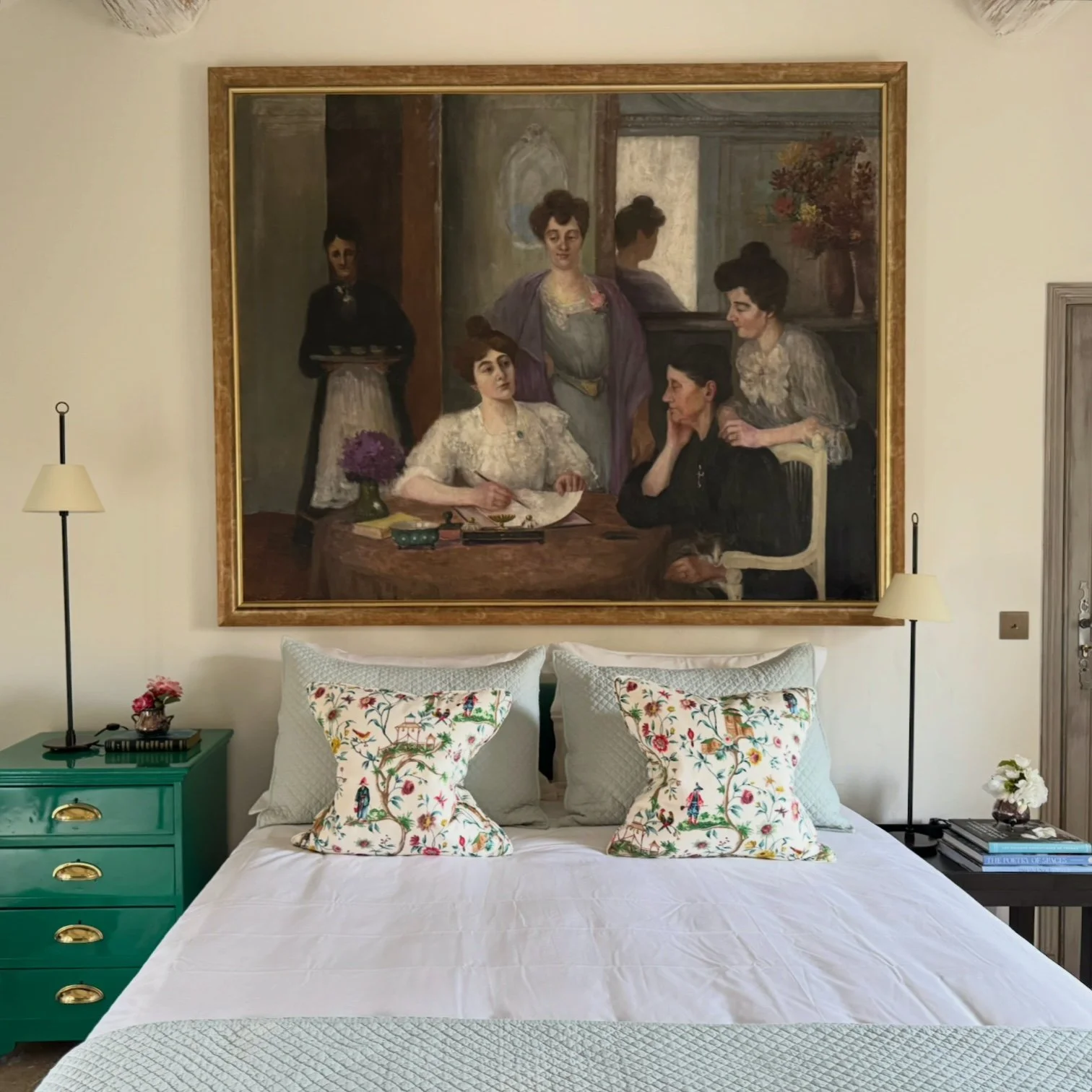 A bedroom with a large framed painting of five women hanging above the bed. The bed has white bedding, two decorative pillows , and two plain pillows. Two bedside tables are on either side; the left one is teal with gold handles and a small pink flow