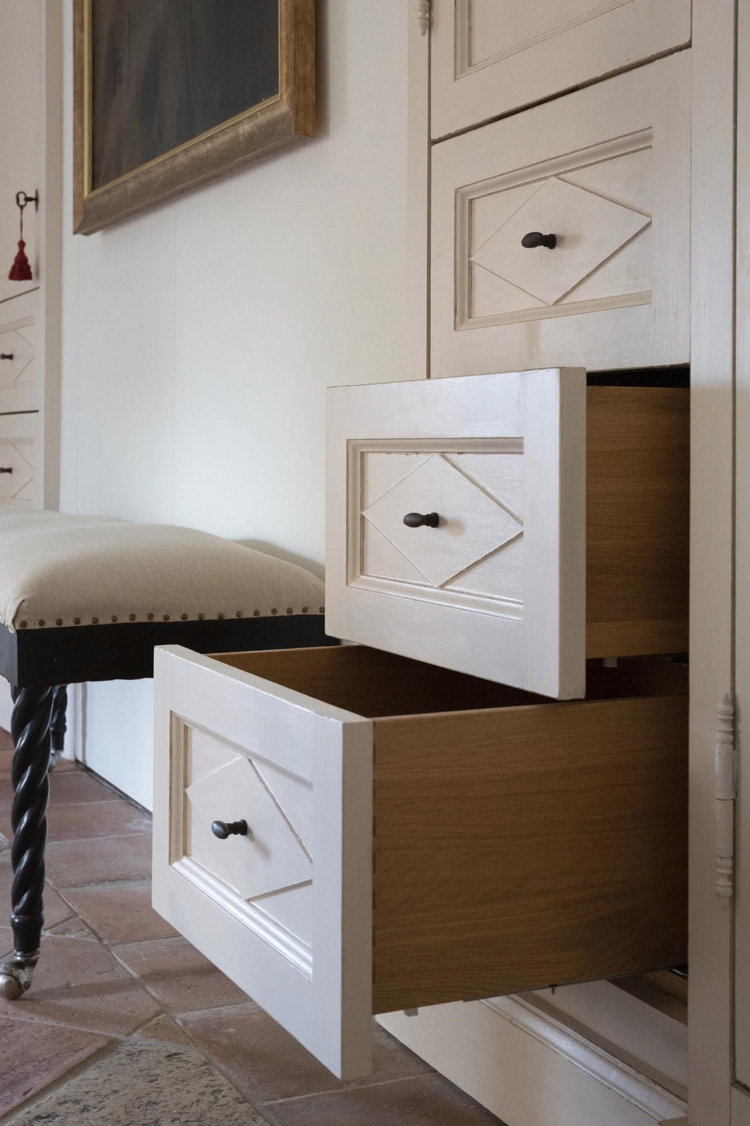 Open drawer in cream-colored wooden furniture next to a beige upholstered chair on a tiled floor.