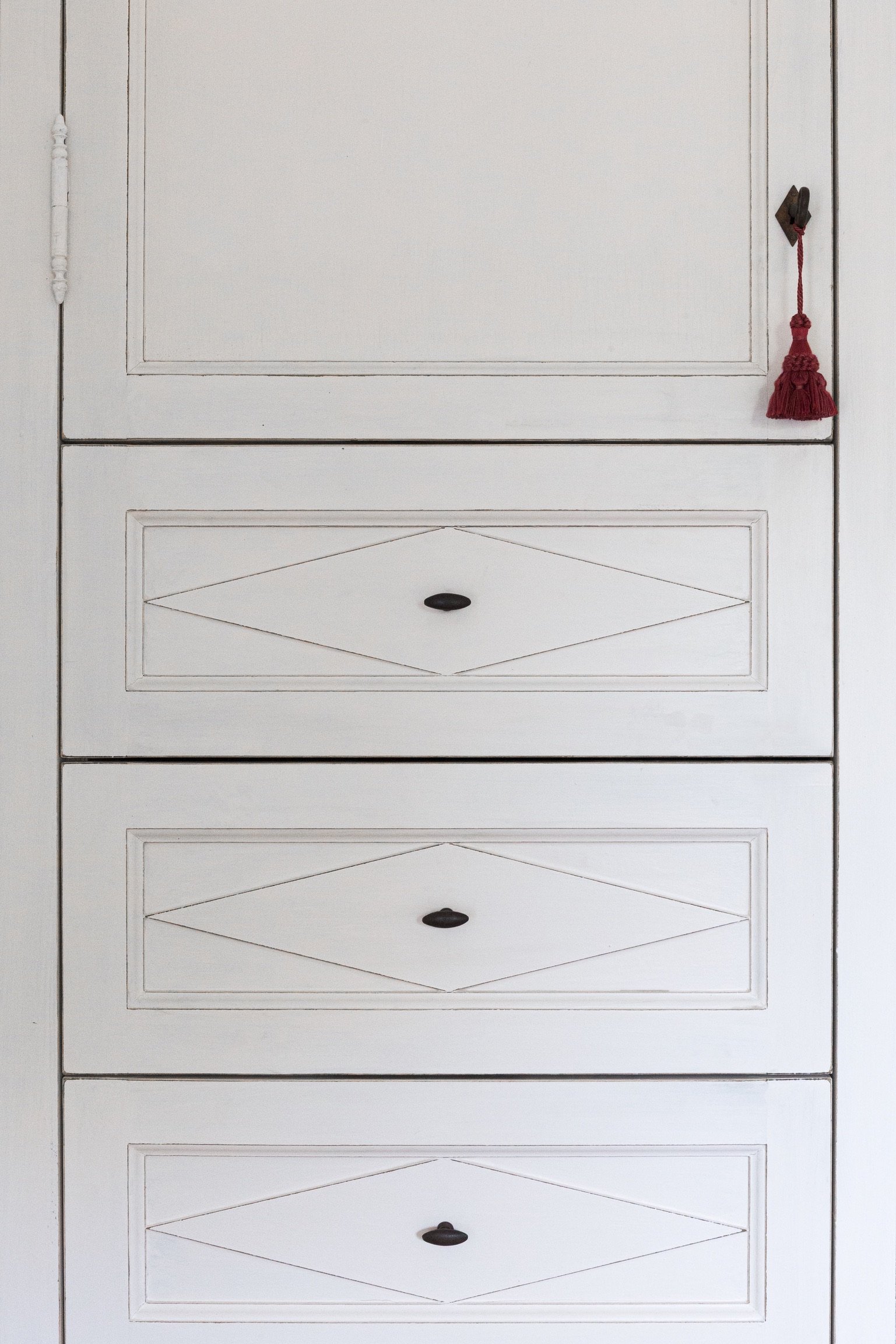 A white wooden dresser with four drawers, each with a black handle, and a decorative carved design. There's a small red tassel hanging from a hook on the top right corner.