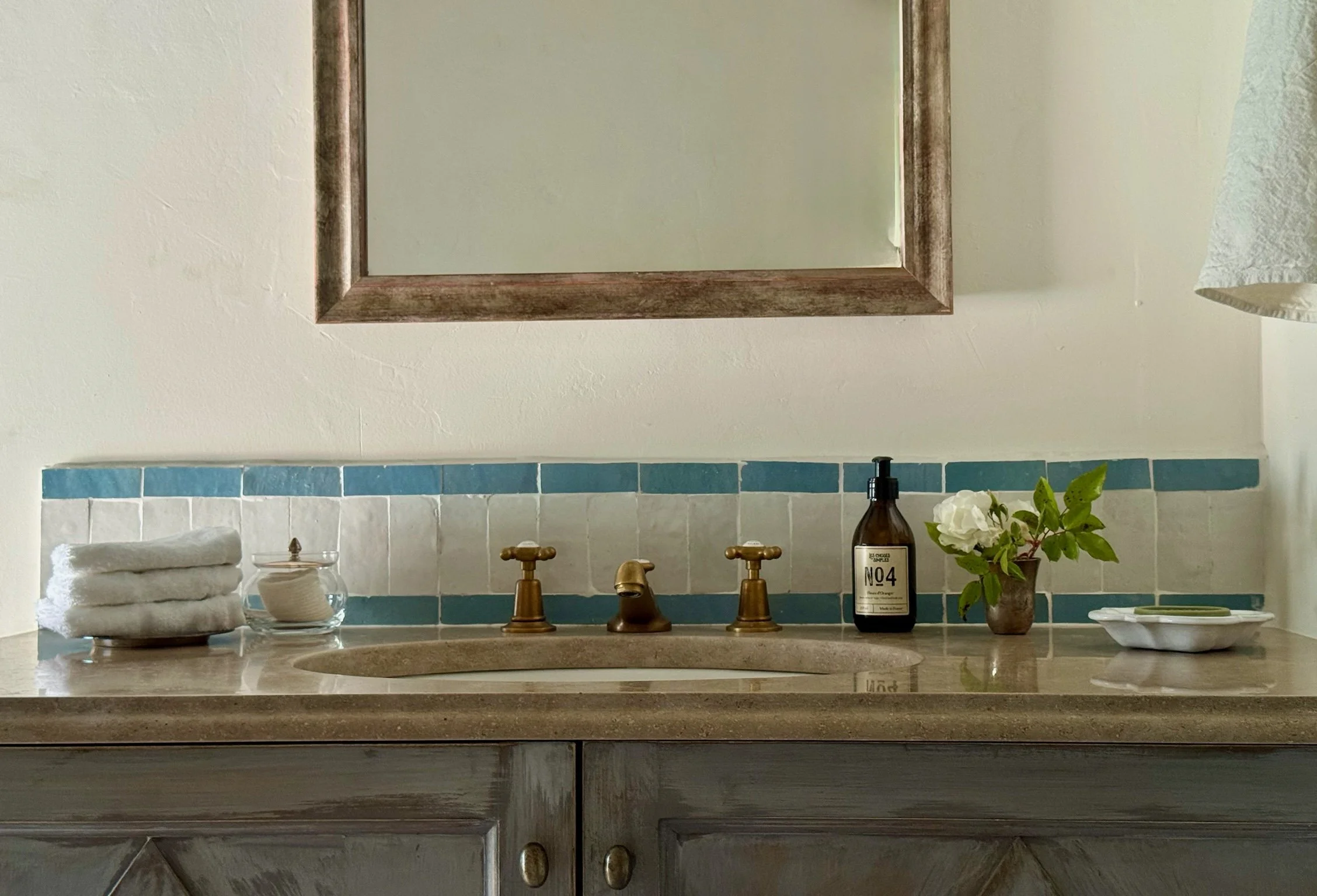 Bathroom sink with stacked white towels, soap jar, brown soap dispenser, small brown vase with white flowers, and a dish with soap, against a wall with blue and white tiles and a mirror above.