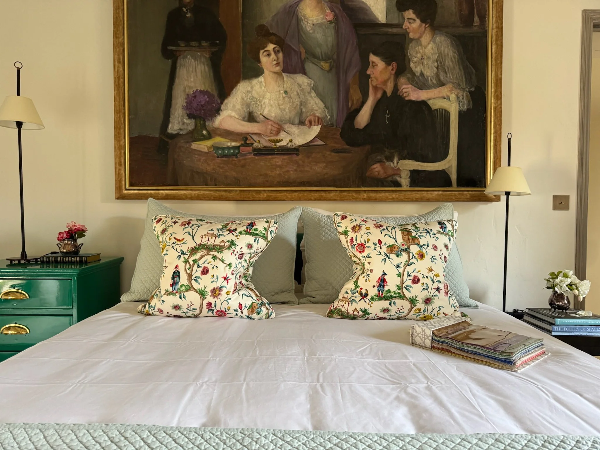 A bedroom with a bed featuring decorative pillows with floral and bird patterns. Above the bed is a large framed painting of four women sitting and standing around a table. On each side of the bed are nightstands with lamps, one green with a small po